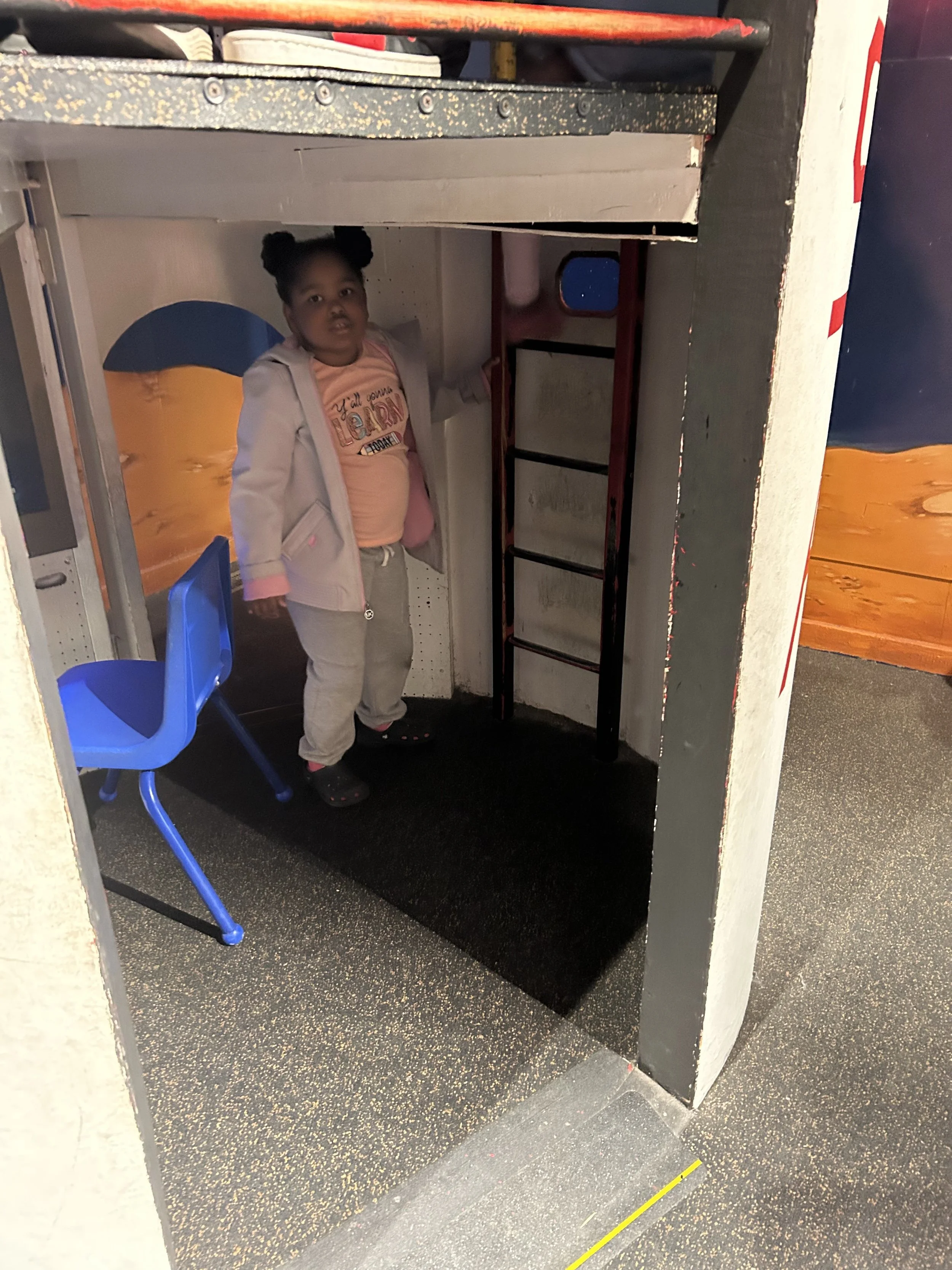 A young girl with two buns hairstyle, wearing a peach shirt with writing, gray sweatpants, and a light jacket, standing under a play structure at an indoor playground.