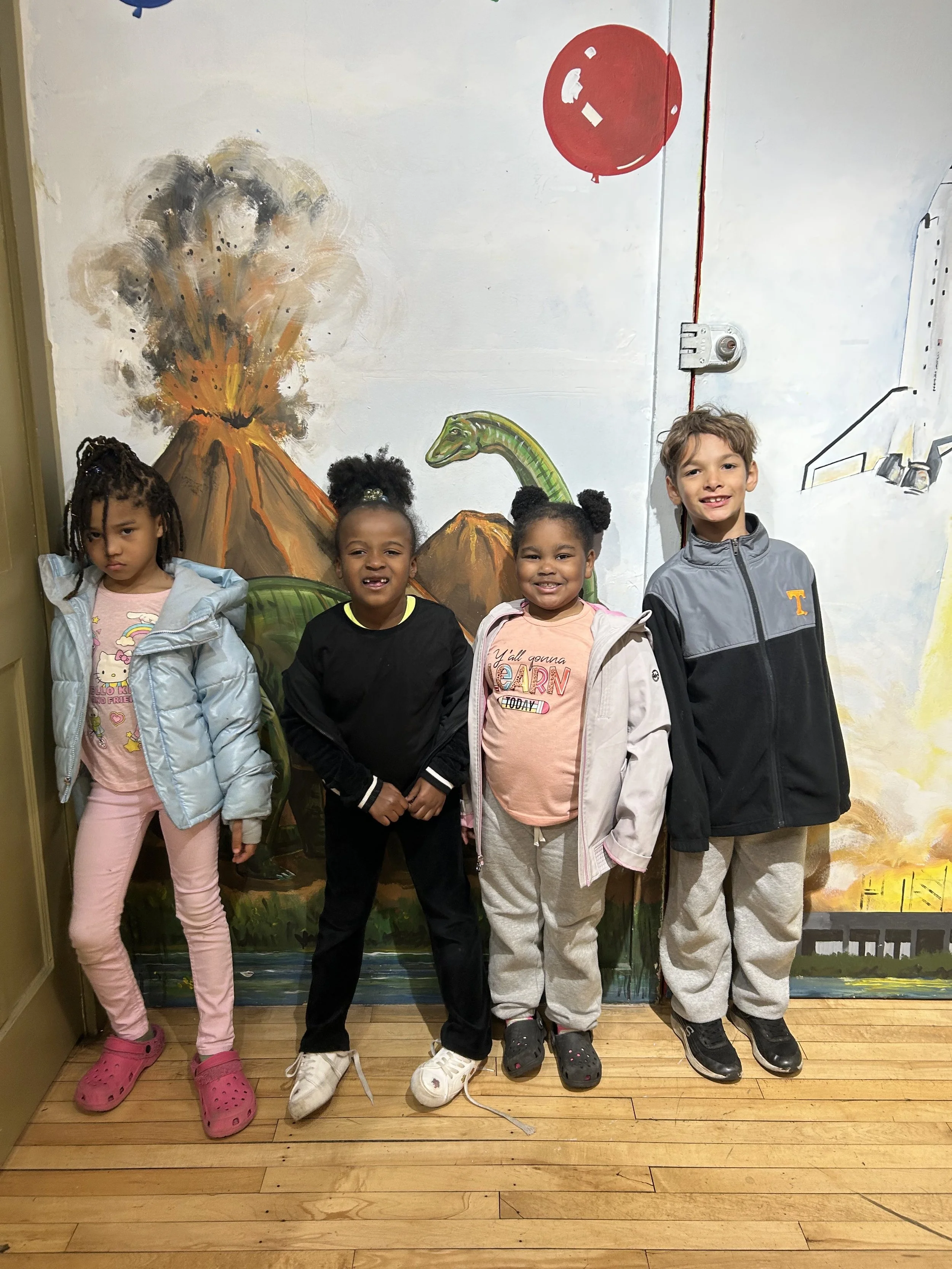 Four children standing in front of a painted mural of a volcano, dinosaurs, and a red balloon, on a wooden floor.