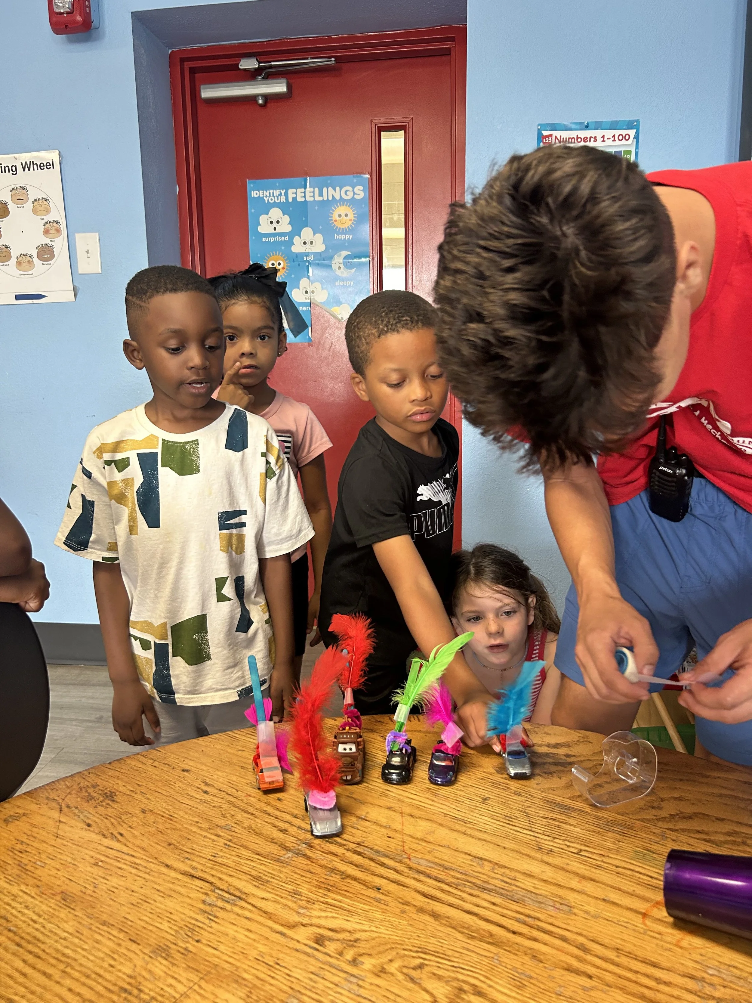 A group of young children gathered around a table, watching an adult prepare a craft project featuring small cars decorated with colorful feathers and other decorations.