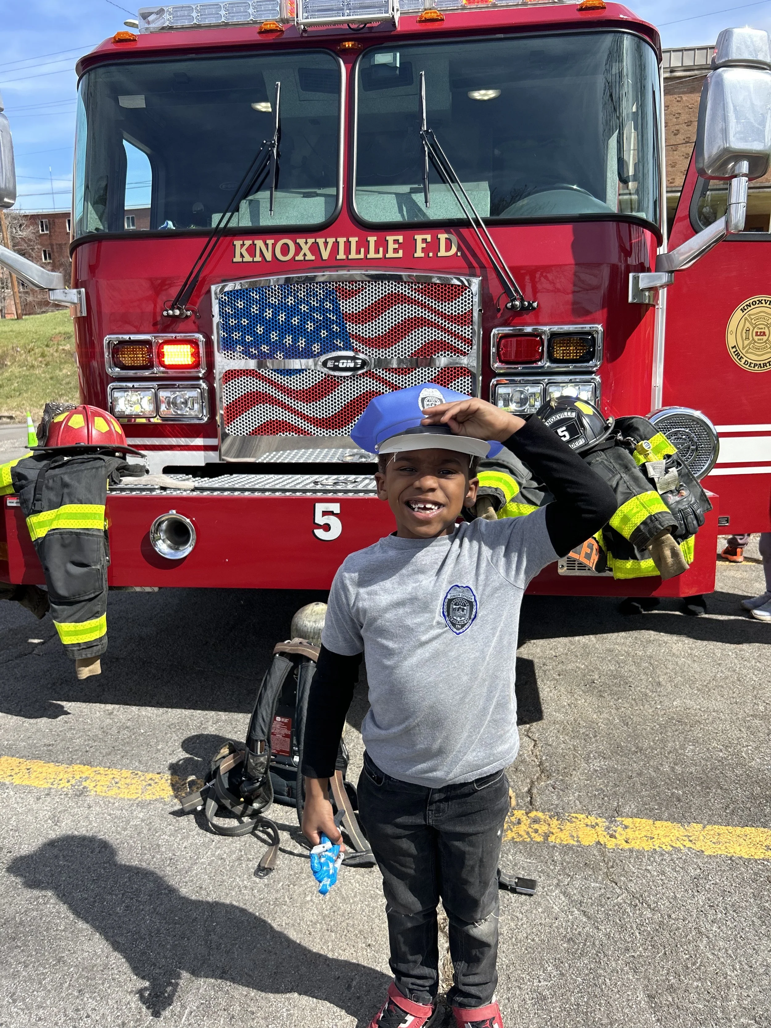 A smiling young boy in a gray shirt with a fire department badge, wearing a police hat and standing in front of a large red fire truck with equipment and hoses. The boy is saluting and holding a small toy.