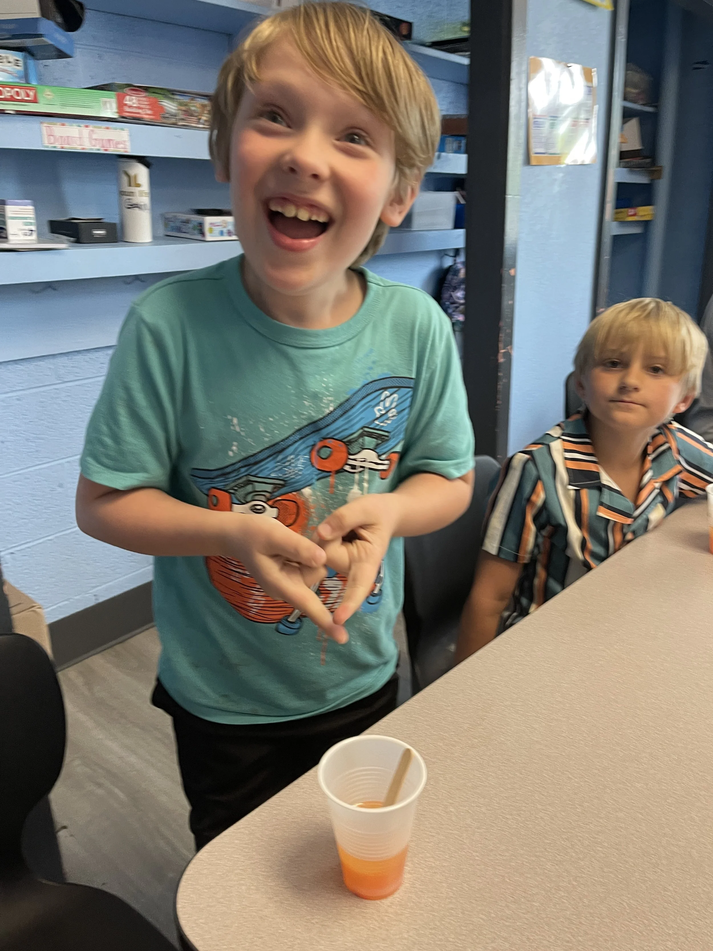 A young boy with light brown hair smiling and playing with a cup of colorful liquid at a table. A second boy with blonde hair and a striped shirt is sitting next to him. The setting appears to be a game or activity room with shelves of board games in