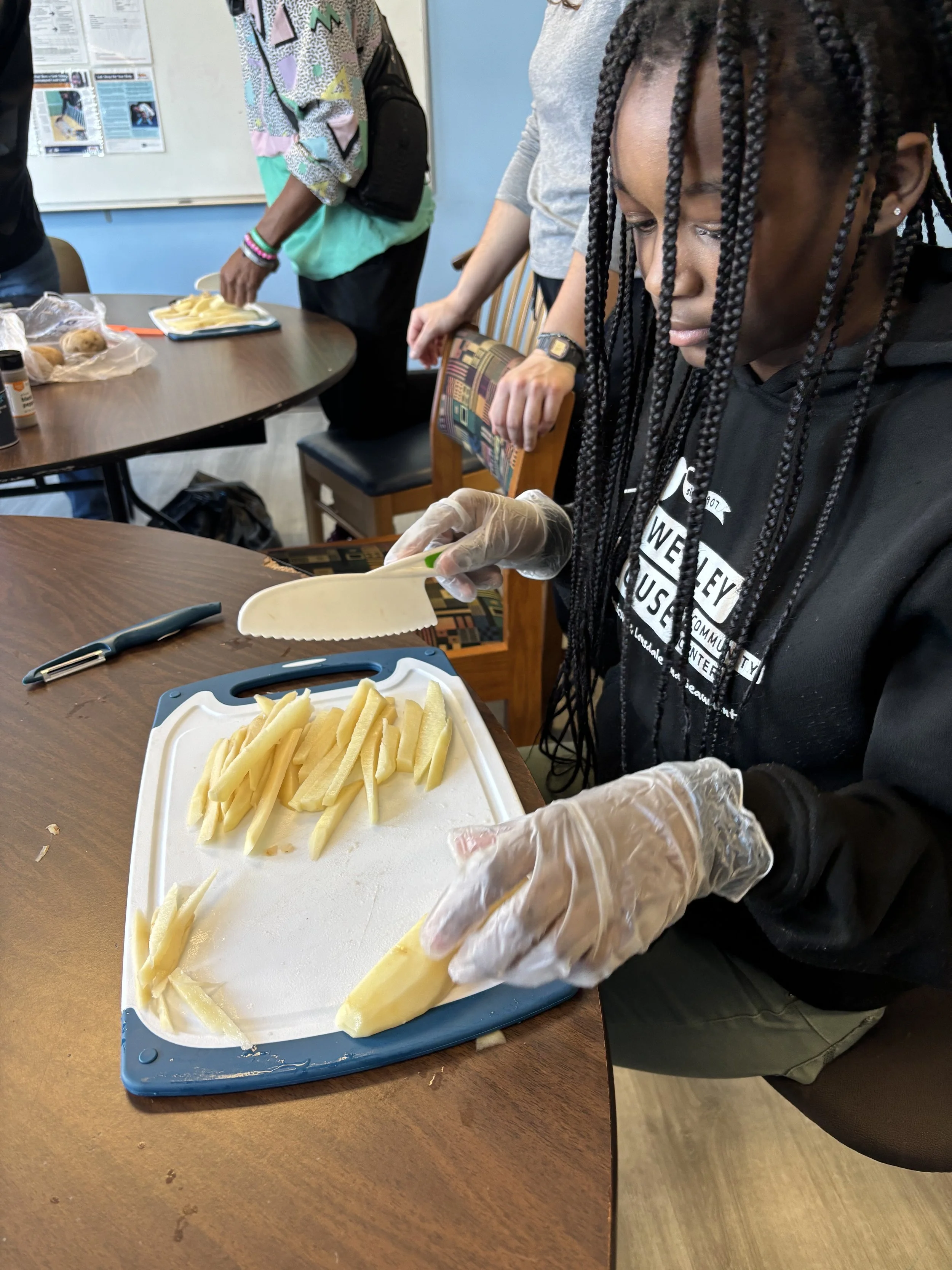 A young woman with braided hair wearing a black hoodie and transparent gloves is slicing a large potato on a white cutting board. There are cut potato strips on the board. She is in a room with other people, some holding trays and standing near a blu
