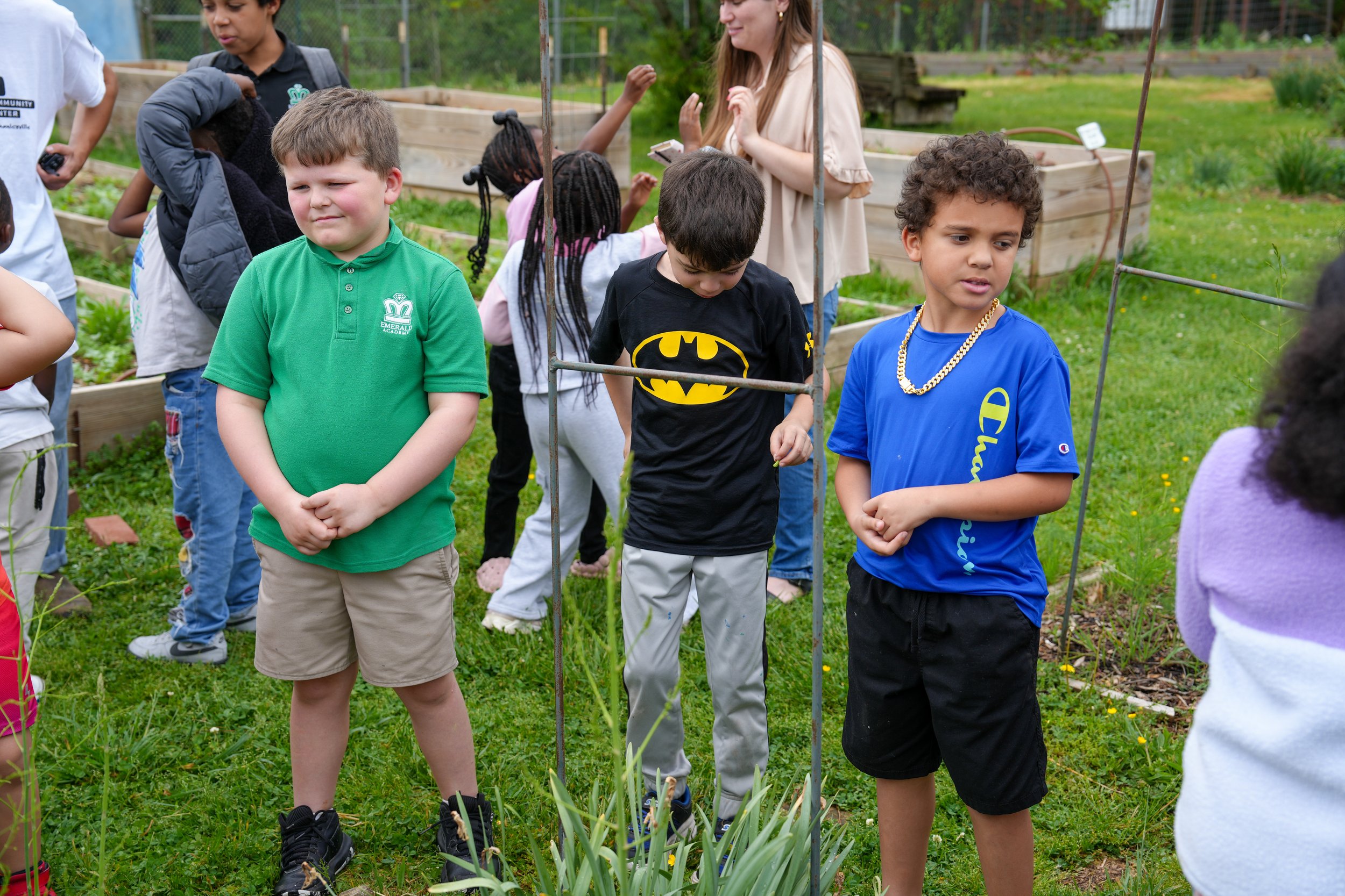 Children standing in a garden, some with their eyes closed, others looking around. They are engaged in an outdoor activity, surrounded by green grass, plants, and garden beds.