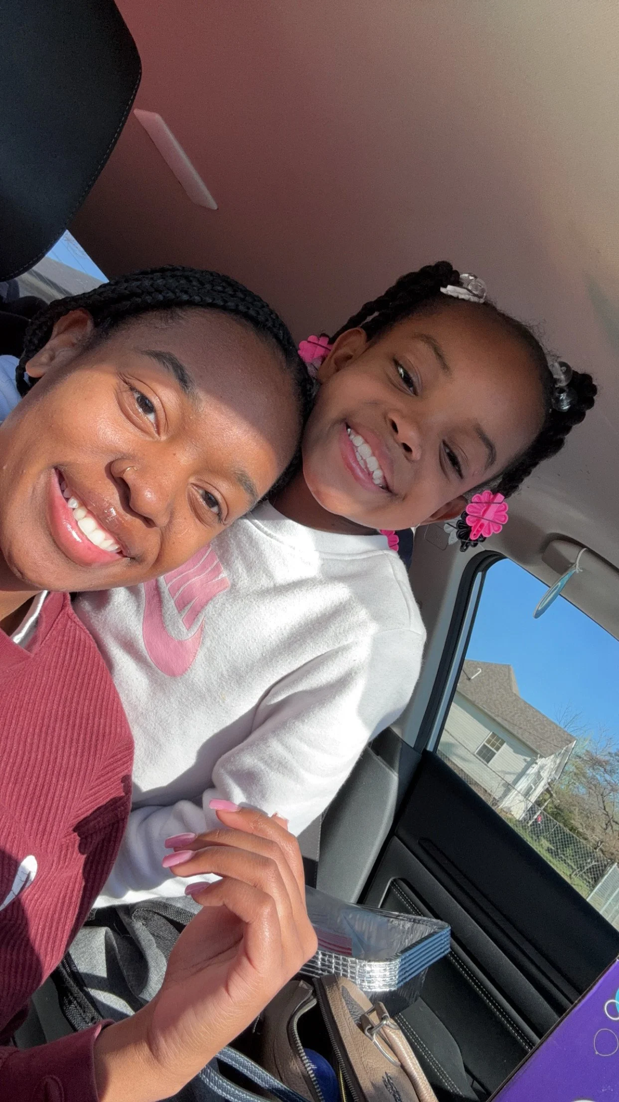 A young girl and an adult woman smiling and posing for a selfie inside a vehicle, with a house and blue sky visible through the window.