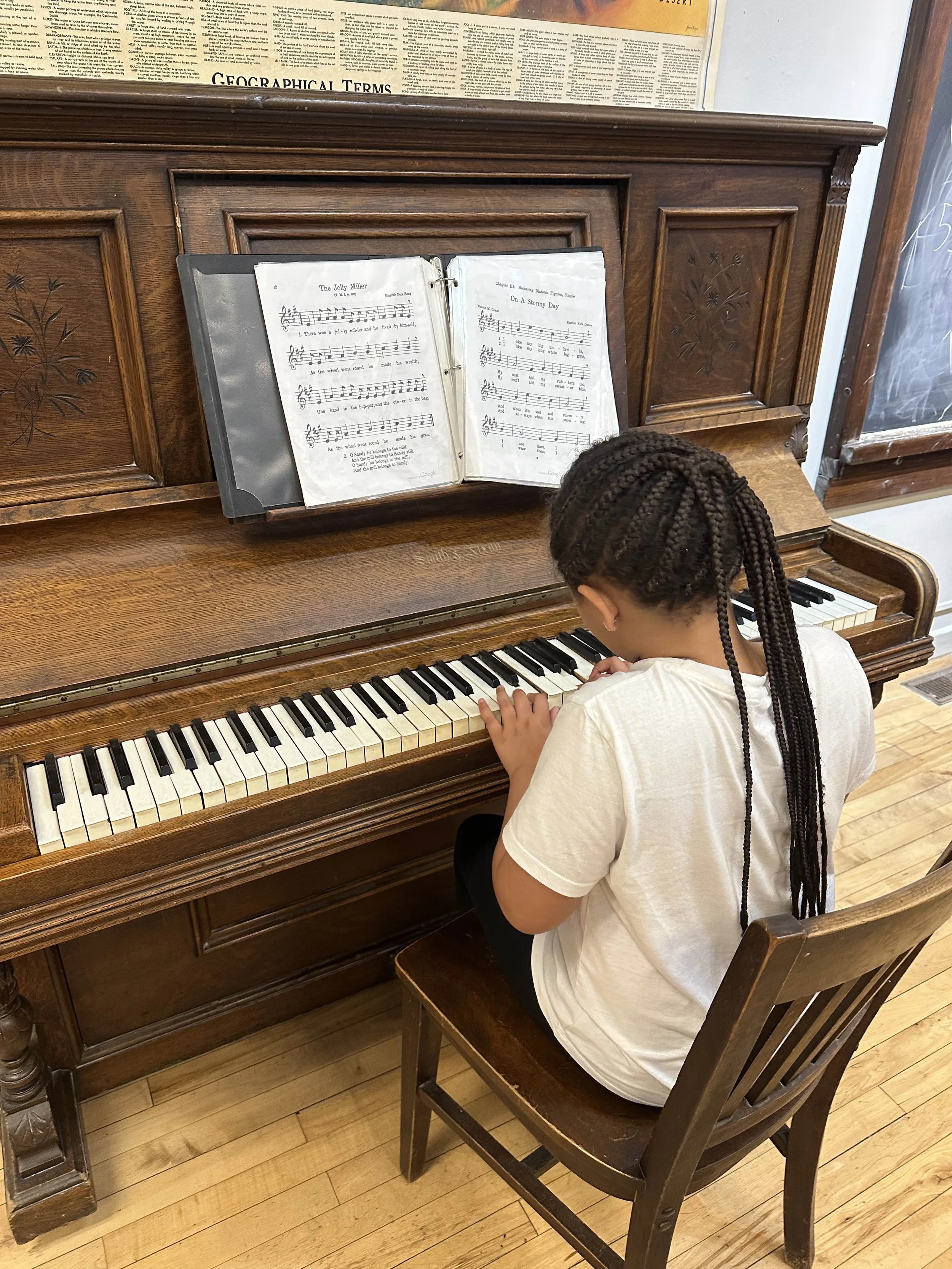 A young girl with braided hair is sitting on a wooden chair playing an upright piano. There is sheet music on the music stand and a large sheet of paper above the piano. The room has hardwood floors and a chalkboard is visible on the right.