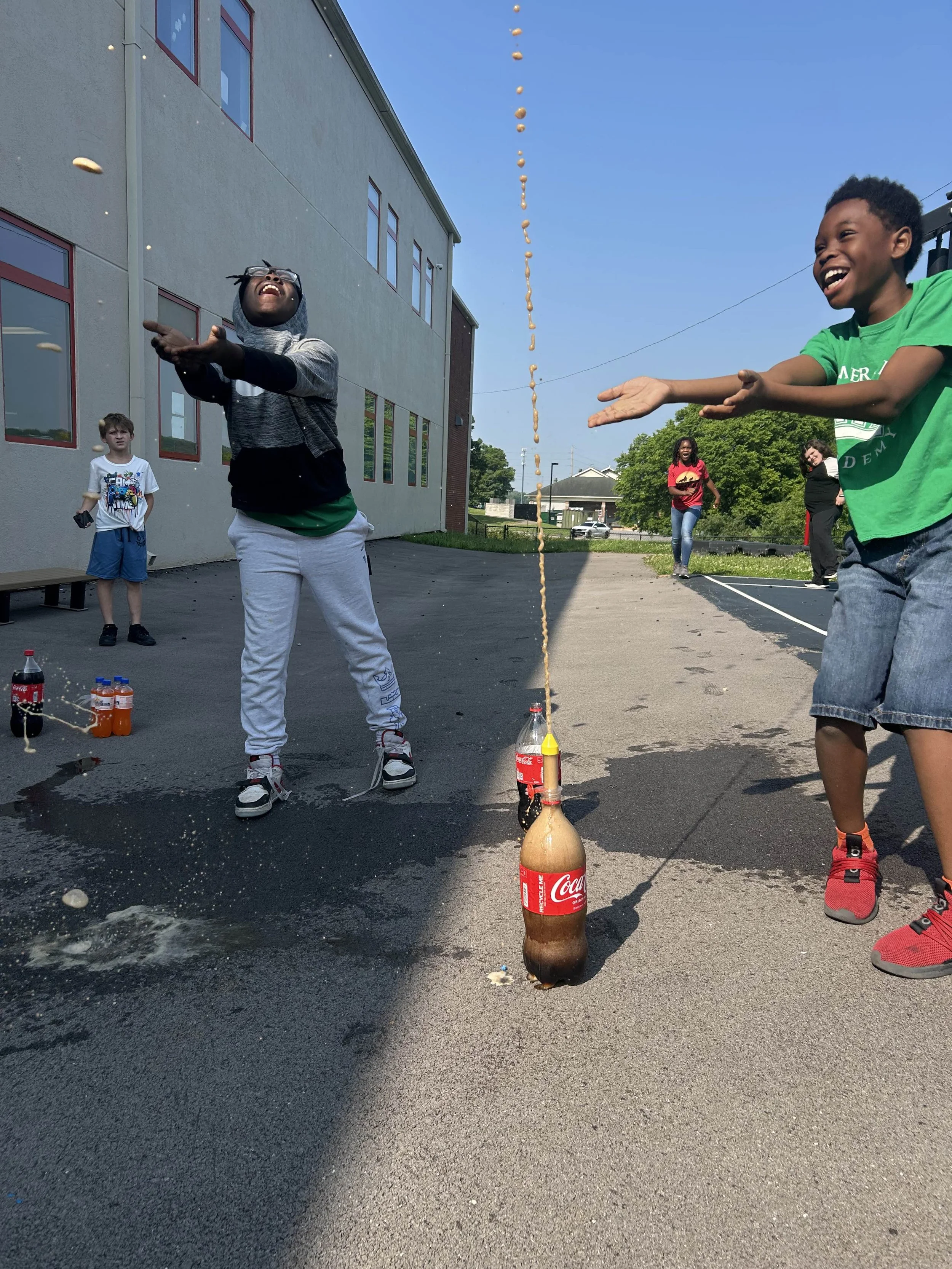 Children playing a game with soda bottles outdoors on a school playground, with a line of small objects dropping from above and kids laughing.