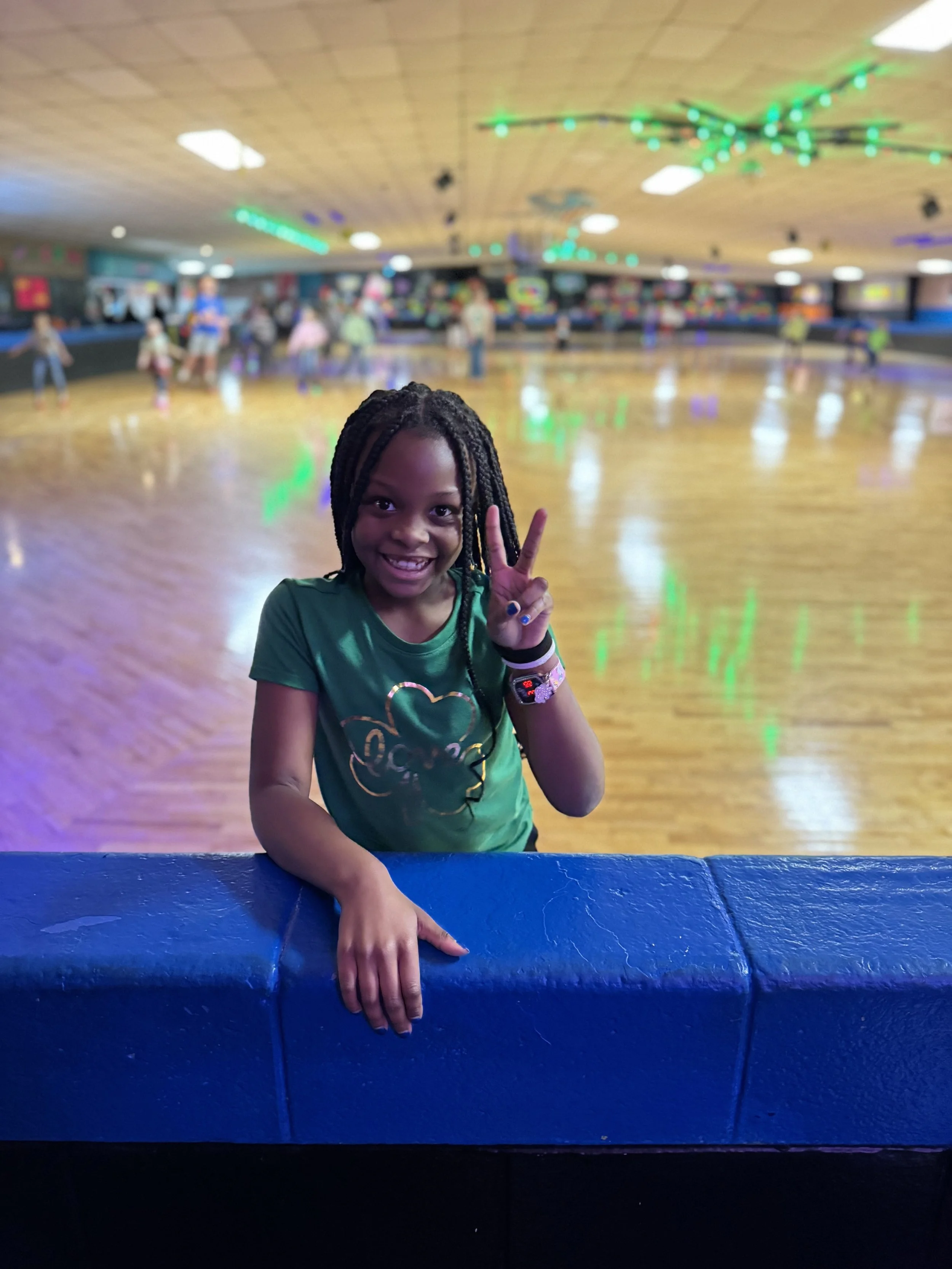 Smiling girl at roller skating rink making peace sign with hand, background features blurred skaters and colorful lights.