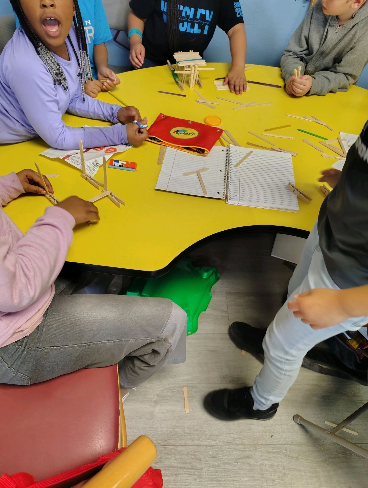Children gathered around a yellow table, building structures with wooden craft sticks.