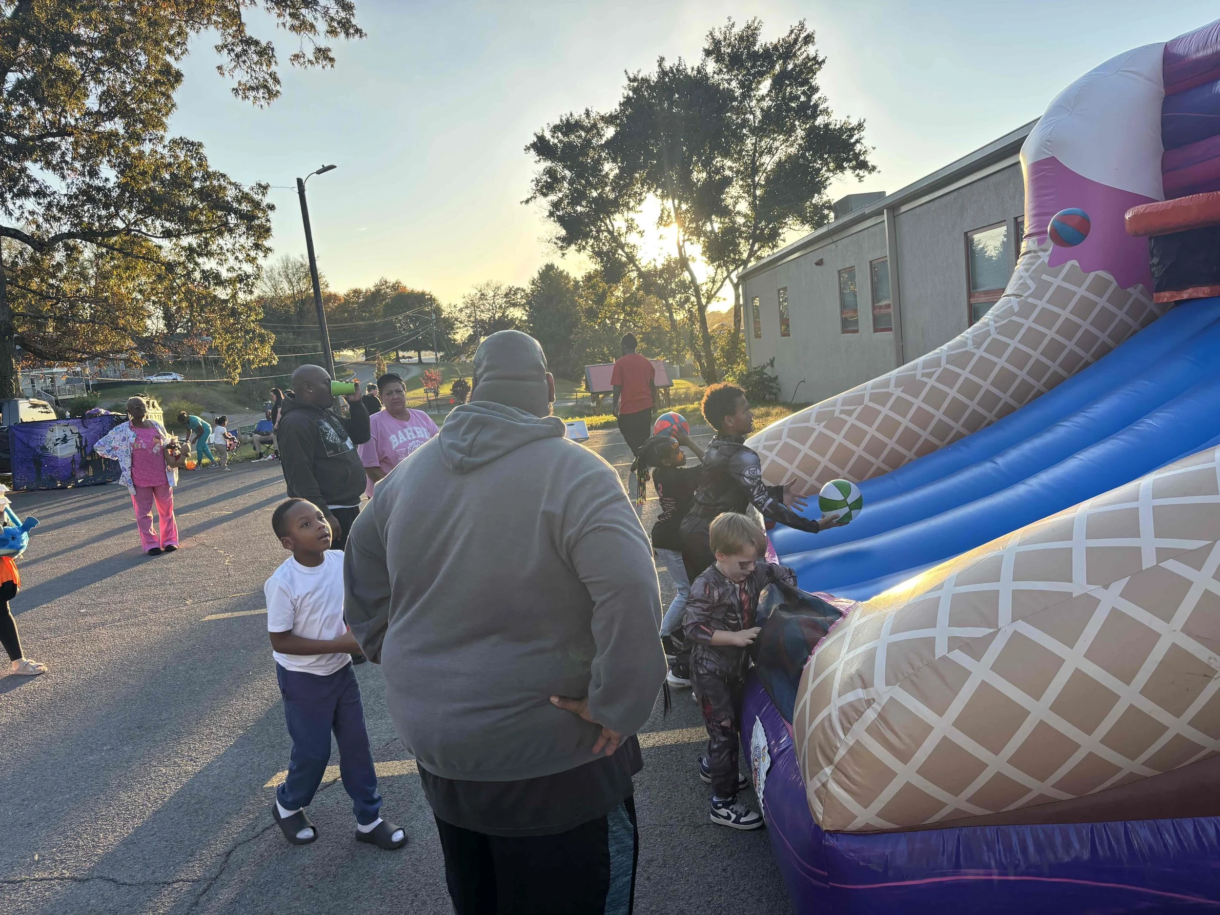 Children and adults gather around an inflatable slide outdoors during late afternoon or early evening. Some children are sliding and playing, while others are waiting or watching. The scene is set in a parking lot with a building, trees, and a clear 