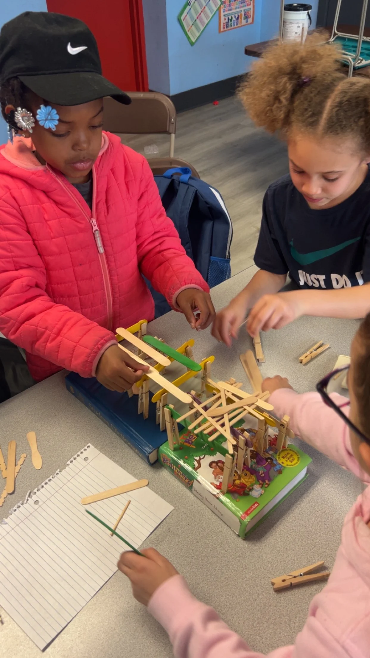 Four children are working together to build a craft project using popsicle sticks on a table in a classroom.