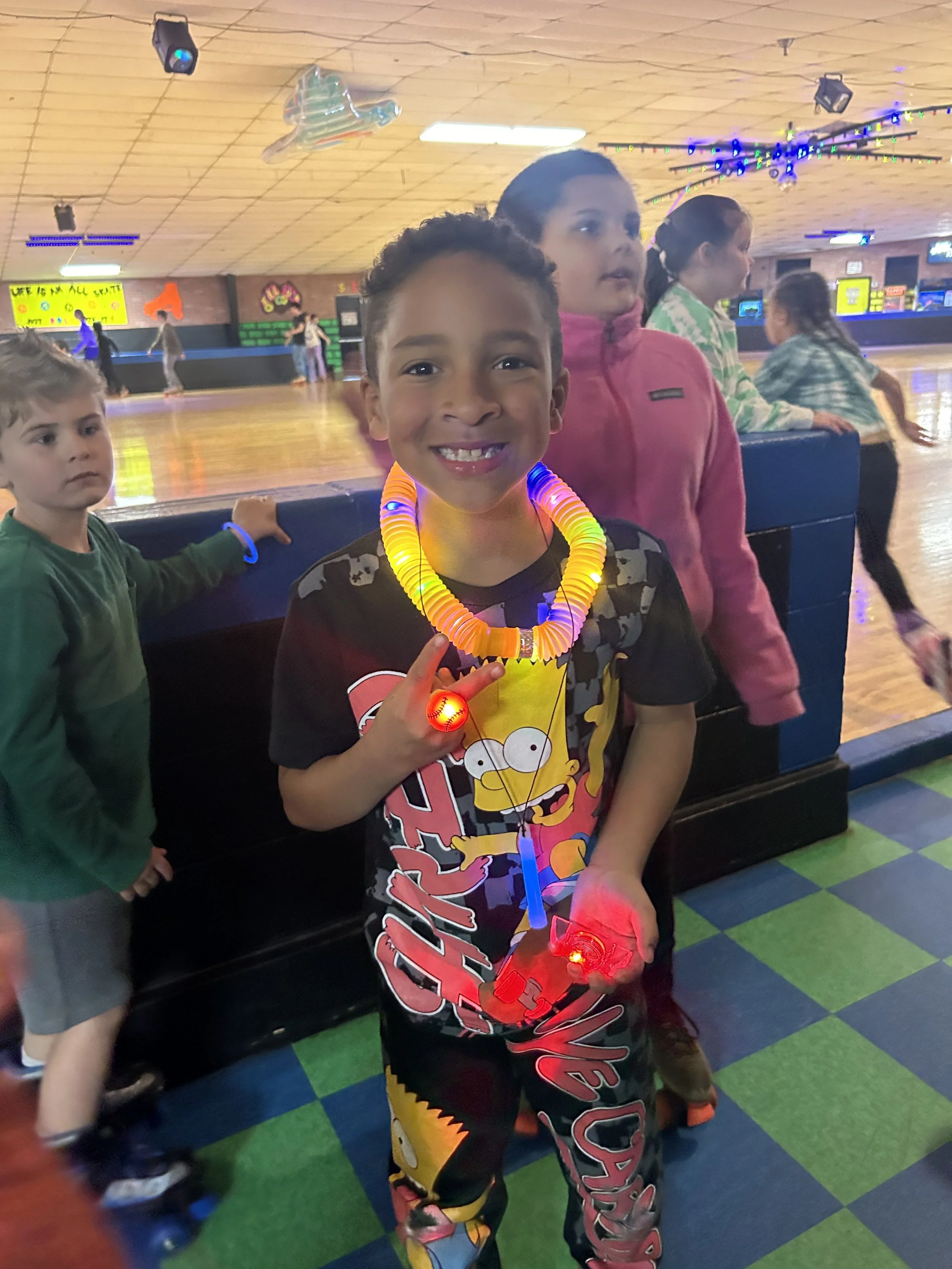 A young boy at an indoor roller skating rink wearing a colorful glowing necklace and holding a red LED device, smiling at the camera. Other children are in the background near the rink.