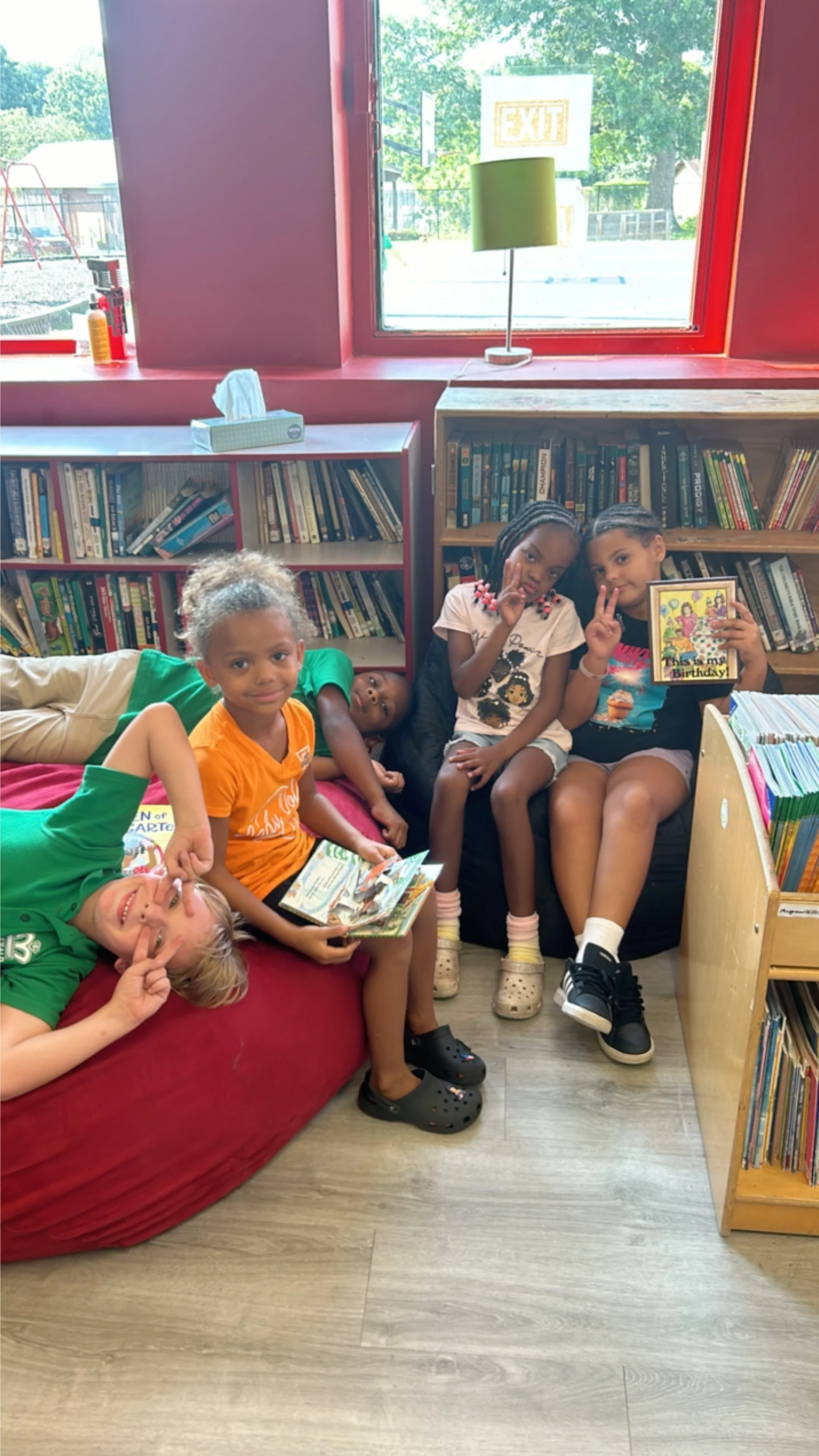 Group of five children sitting and lying on red cushioned seating in a library, holding books and making peace signs, with bookshelves and window in the background.