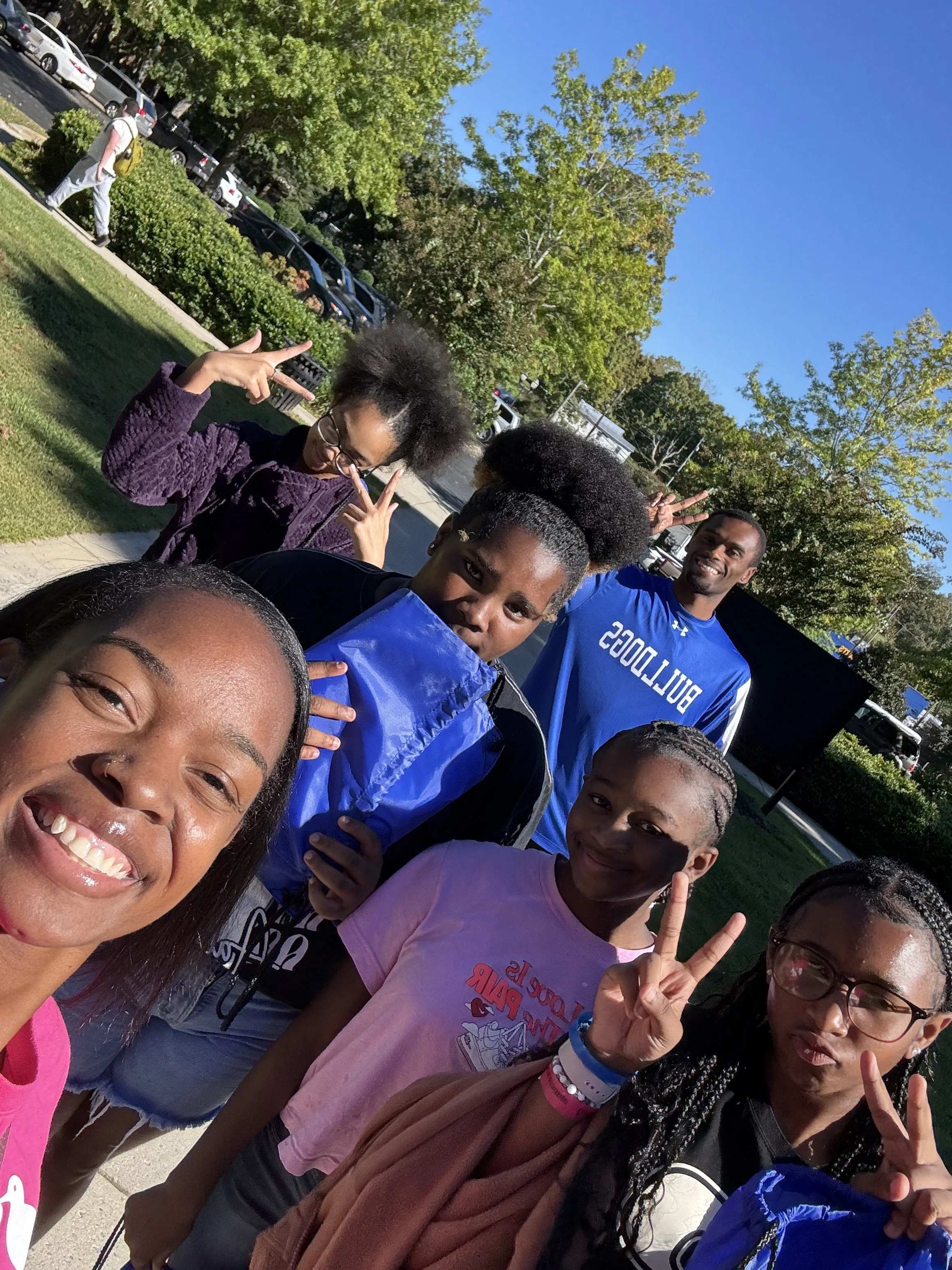 Group of six people smiling and posing for a selfie outdoors on a sunny day, with trees and parked cars in the background.