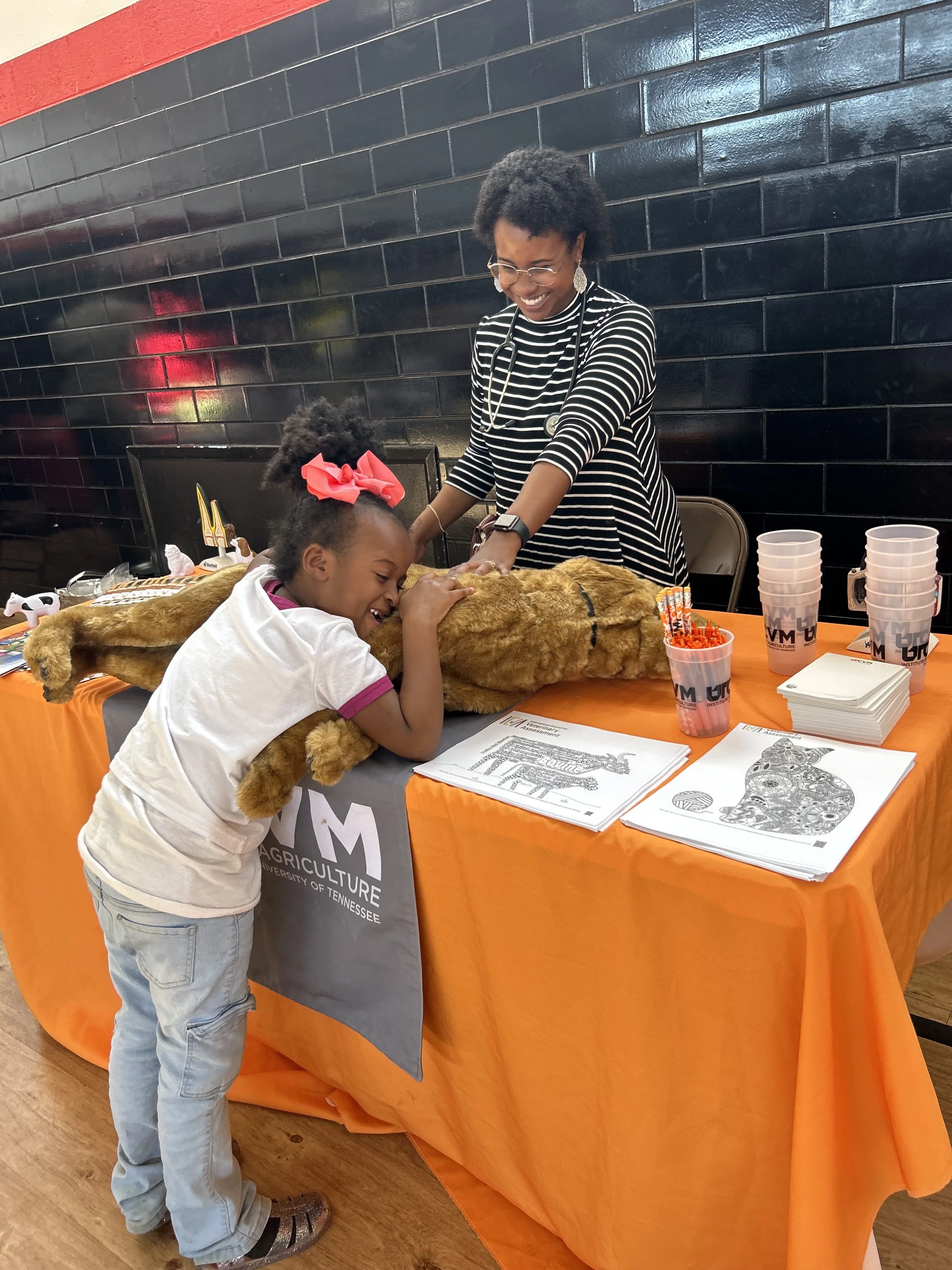 A young girl and an adult woman are petting a live lion lying on an orange table, with the girl smiling and hugging the lion from the side. The table has informational sheets and cups on it, and the background features a black tiled wall.