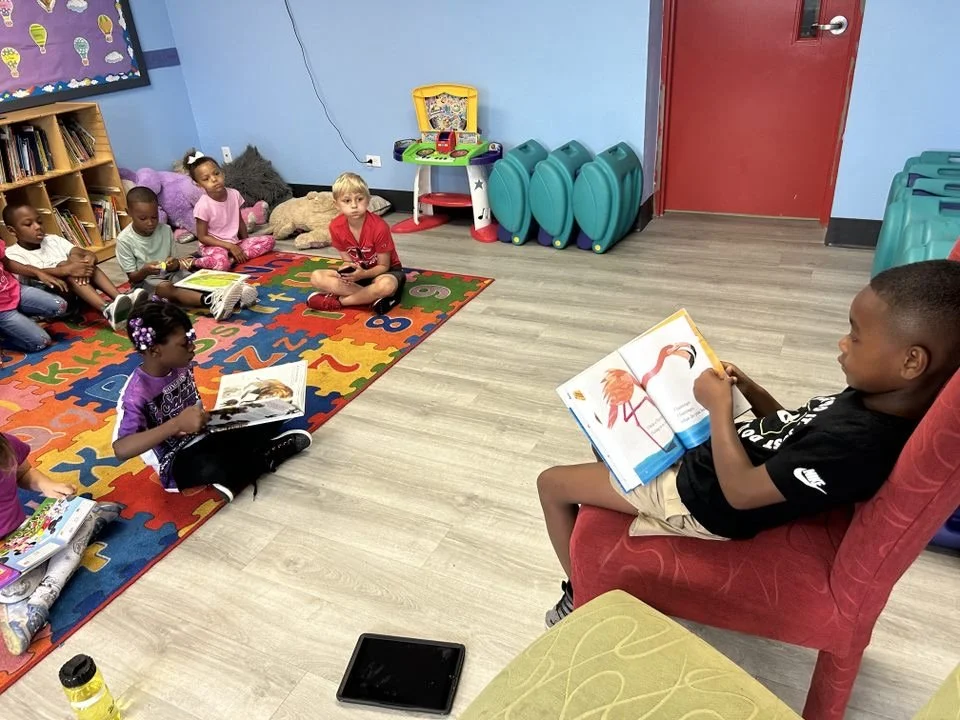 A group of young children sitting on a colorful alphabet rug, listening to a person reading a picture book in a classroom with bookshelves, stuffed animals, and blue storage containers.