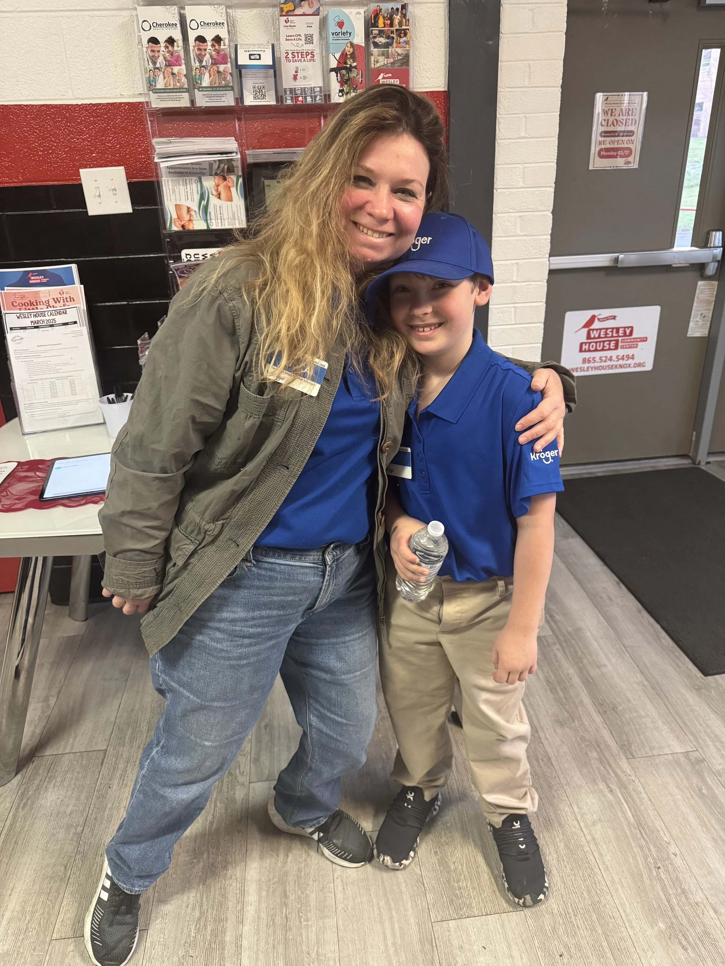 A woman and a young boy wearing Kroger uniforms are smiling and hugging inside a store near the entrance. The woman has long blond hair and is wearing a gray jacket and jeans. The boy is holding a water bottle, wears beige pants, a blue cap, and a bl