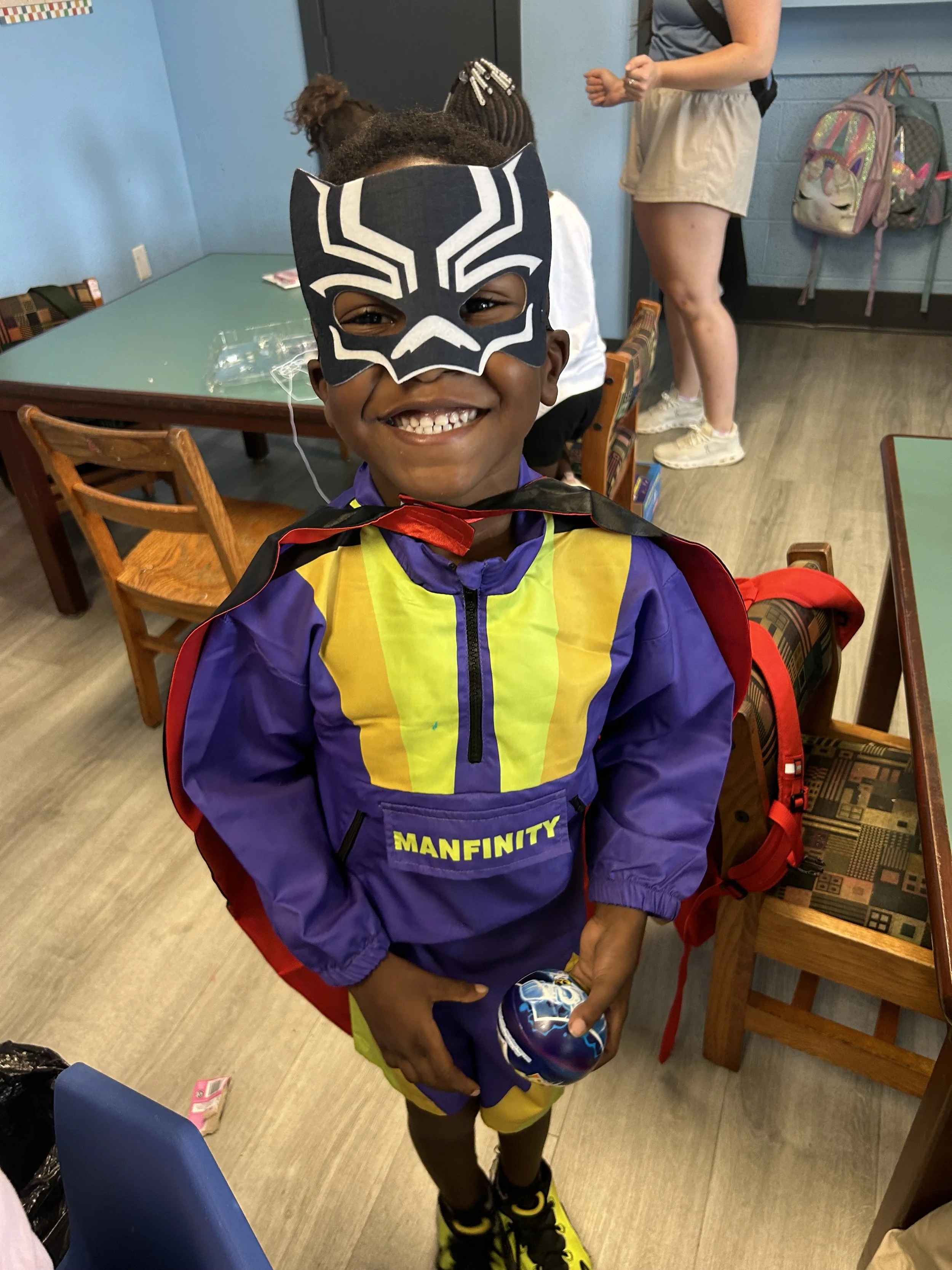 A young boy dressed in a colorful 'Manfinity' superhero costume with a yellow, purple, and black jacket and matching shorts, smiling and holding a small ball, in a room with wooden chairs, a table, and backpacks.