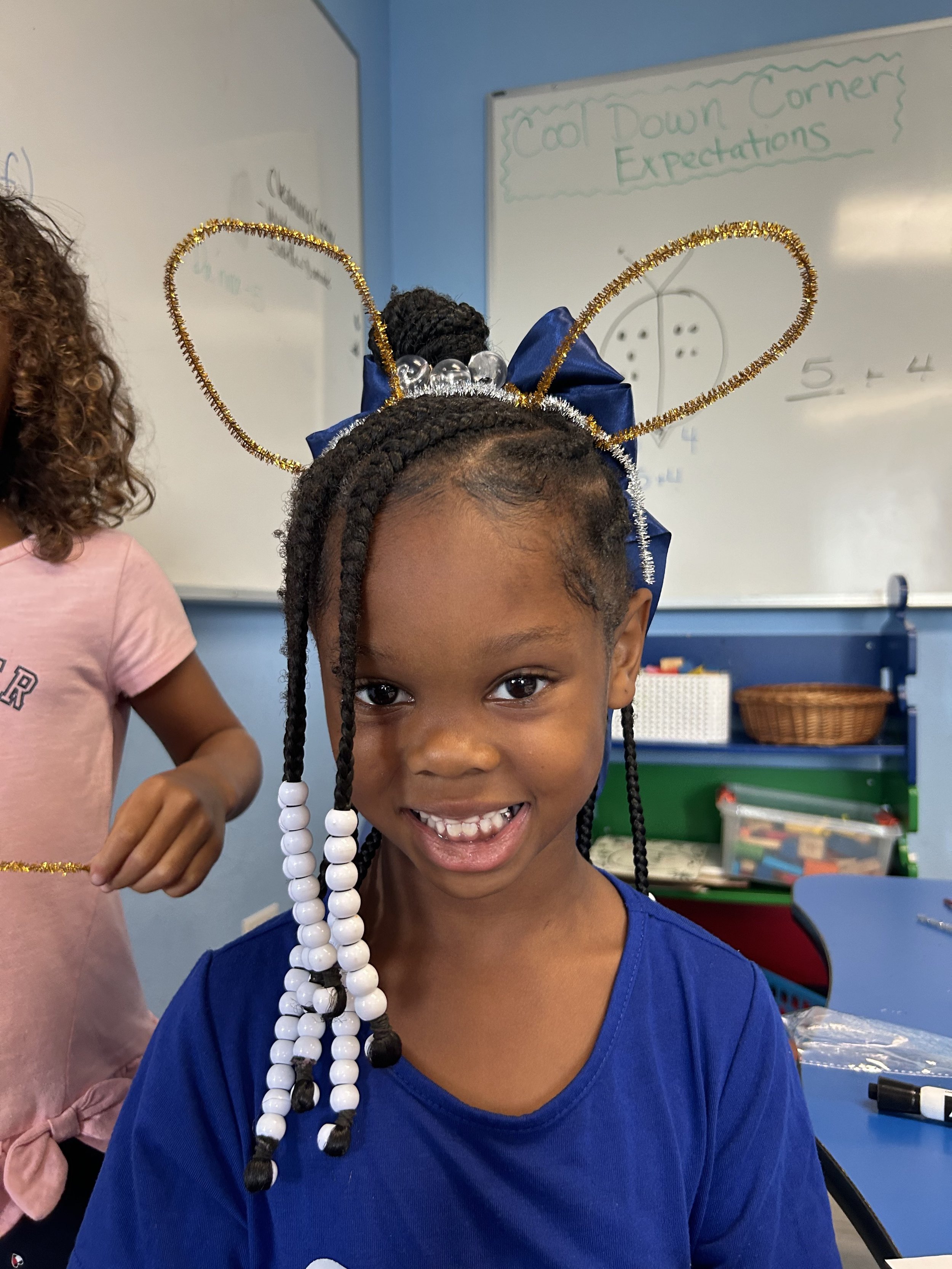 A young girl smiling in a classroom decorated for a celebration, wearing a headband with gold glitter loops, a large blue bow, and black and white beads in her braided hair.