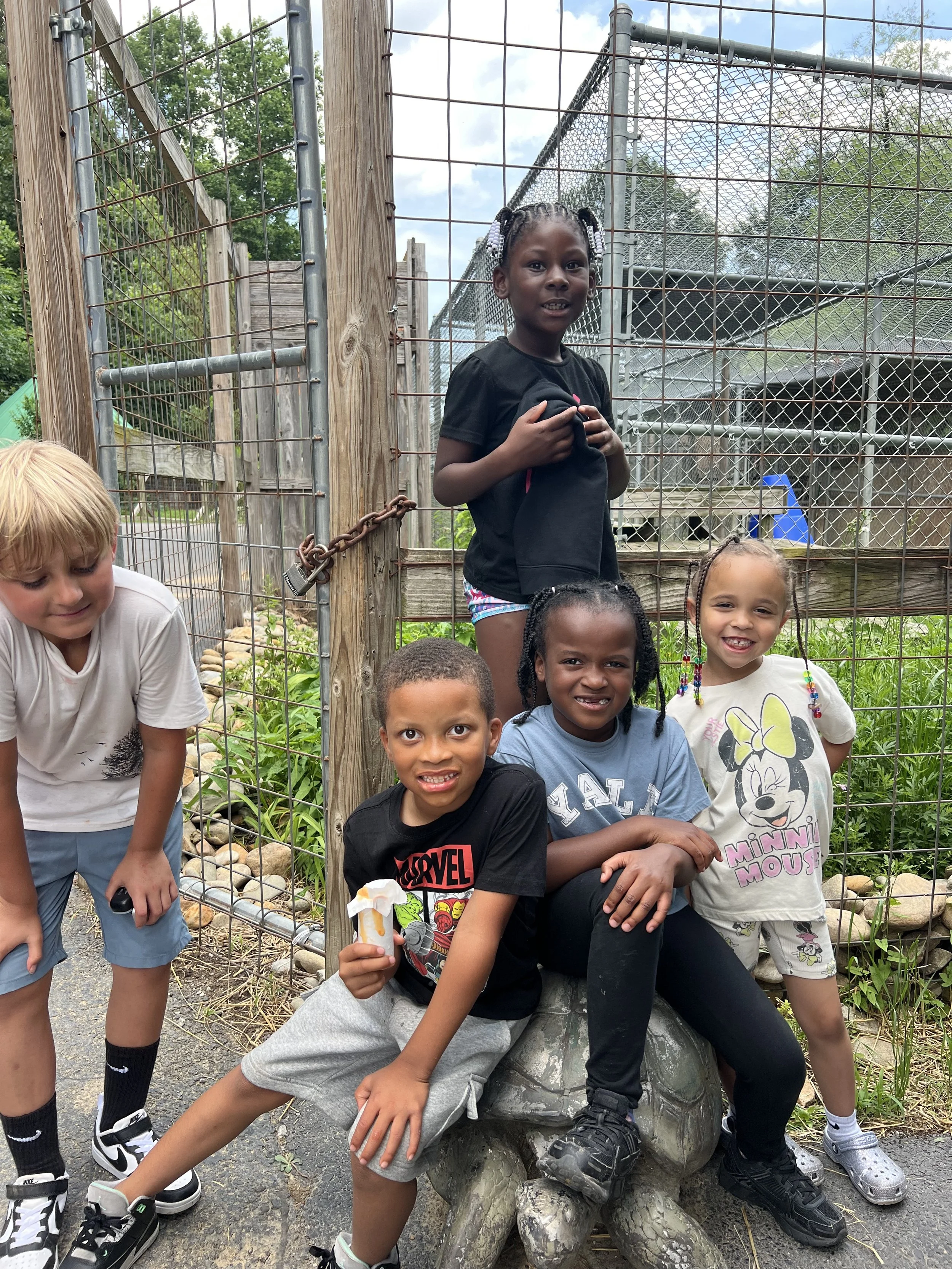Group of five children posing outside near a turtle sculpture, with a fenced enclosure in the background.