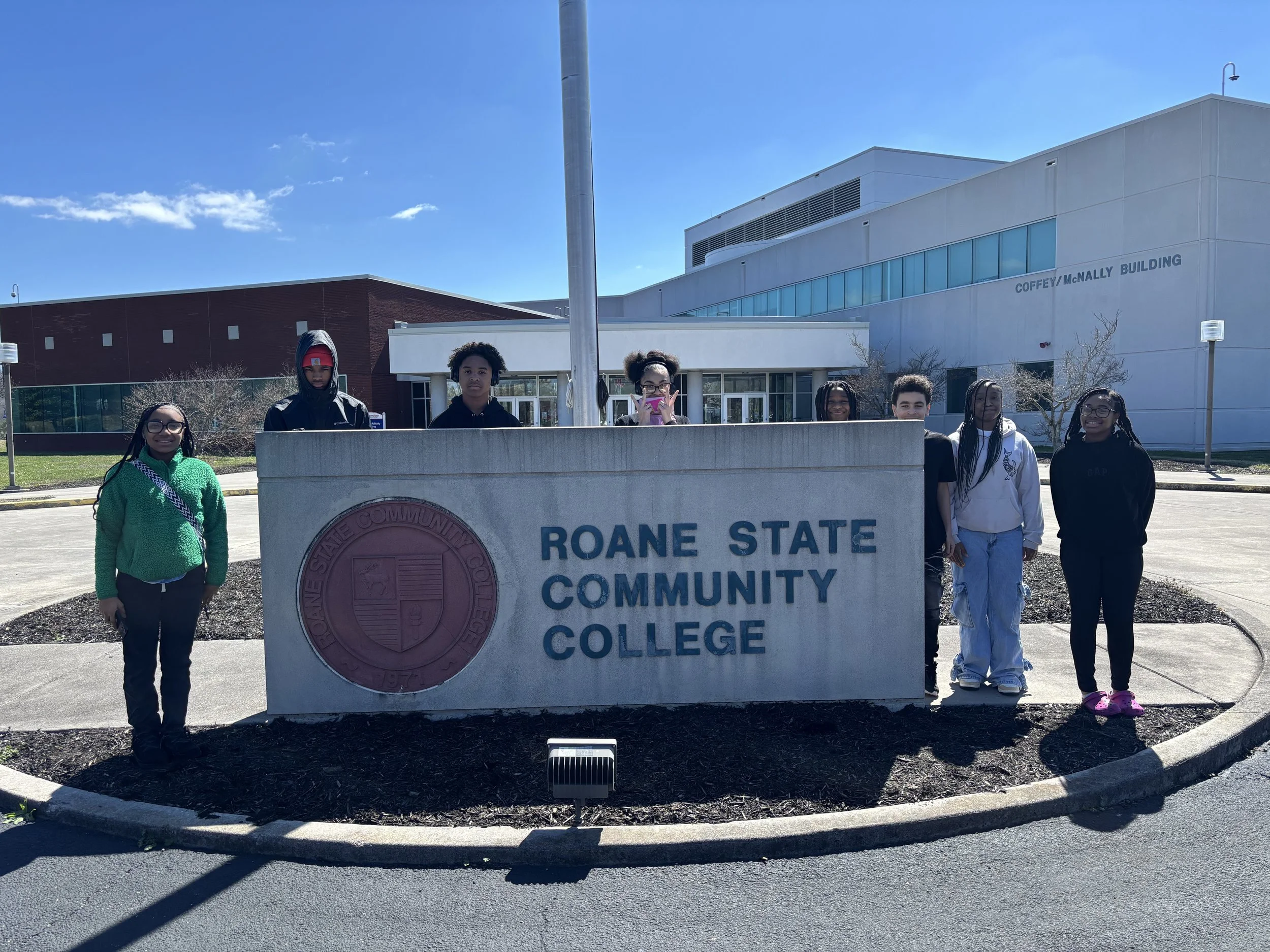 Group of eight students standing behind a sign for Roane State Community College on a sunny day, outside the college building.