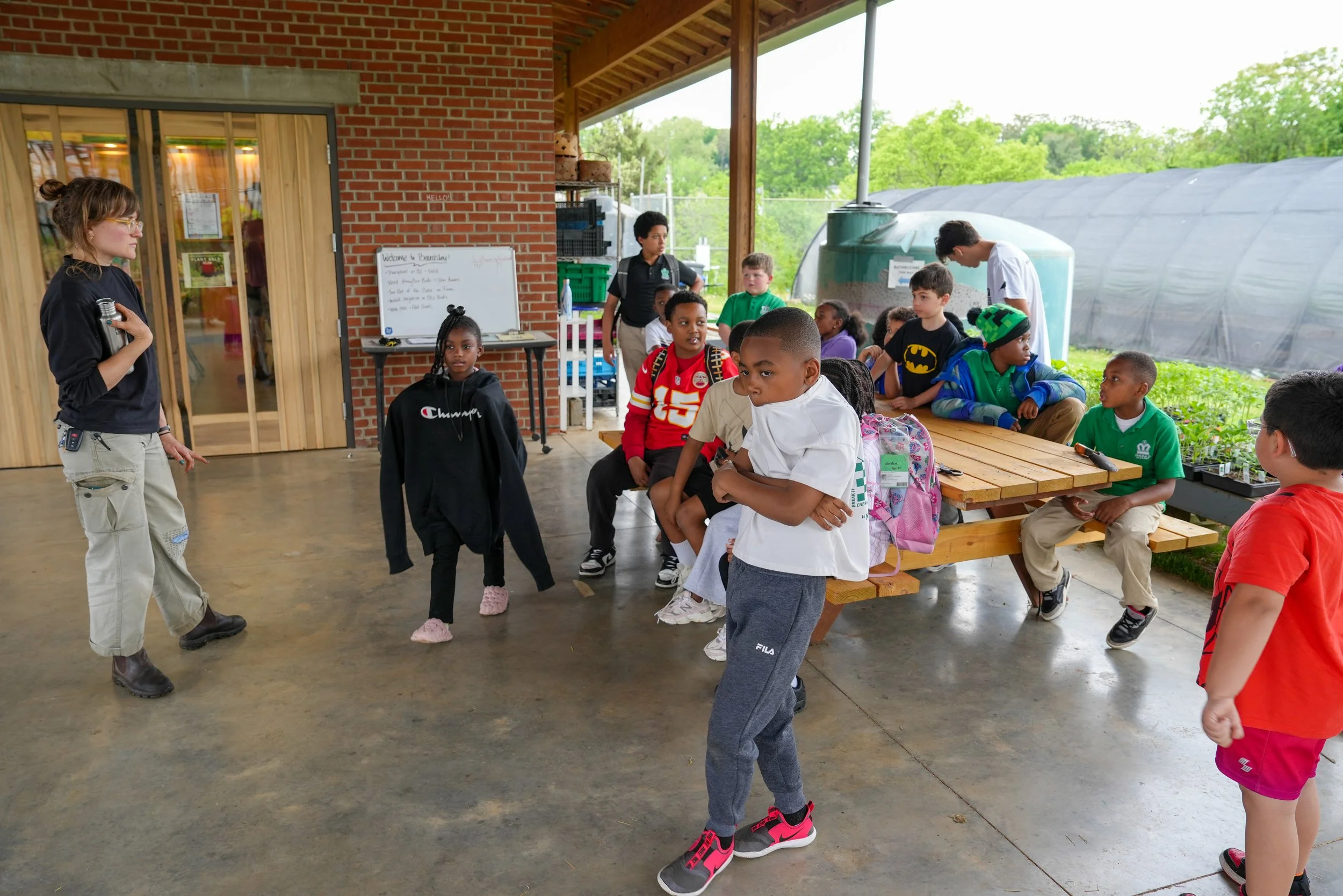 Group of children sitting and standing on a covered outdoor patio, listening to a woman holding a water bottle who appears to be instructing them. Some children are seated at a picnic table, while others stand nearby. There is a whiteboard and some p