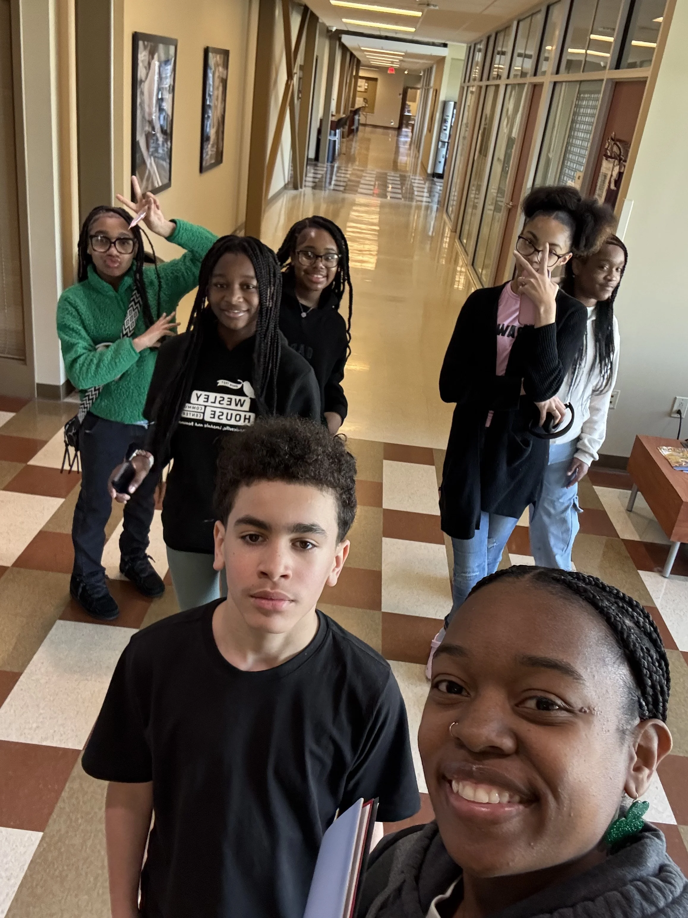 A selfie of a group of young people in a hallway, some making fun and peace signs, with a checkered floor and door panels.