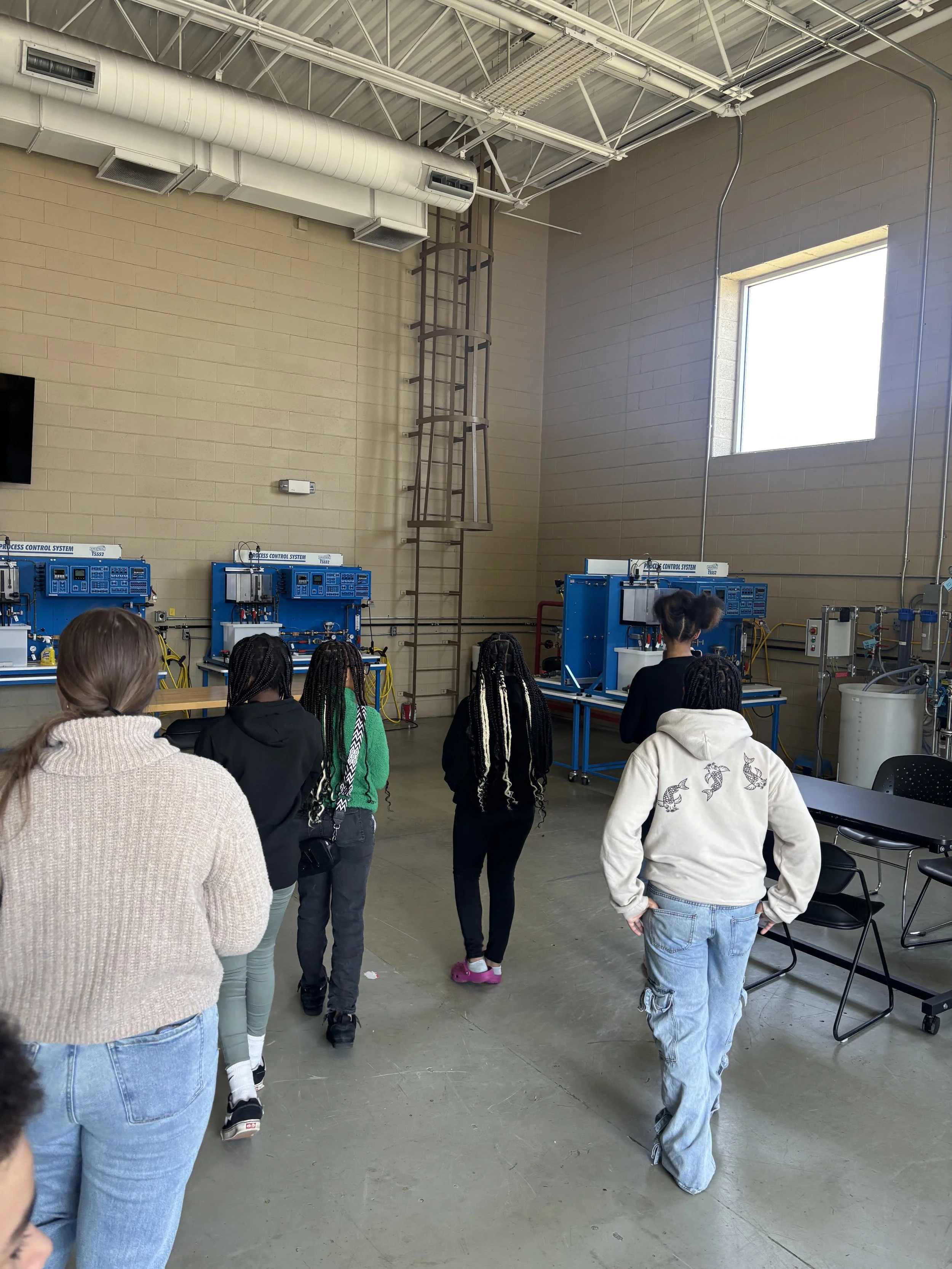 A group of people standing in a science or technology classroom, facing away, observing equipment on tables along the wall. The room has a high ceiling with exposed ductwork and a large window letting in natural light.