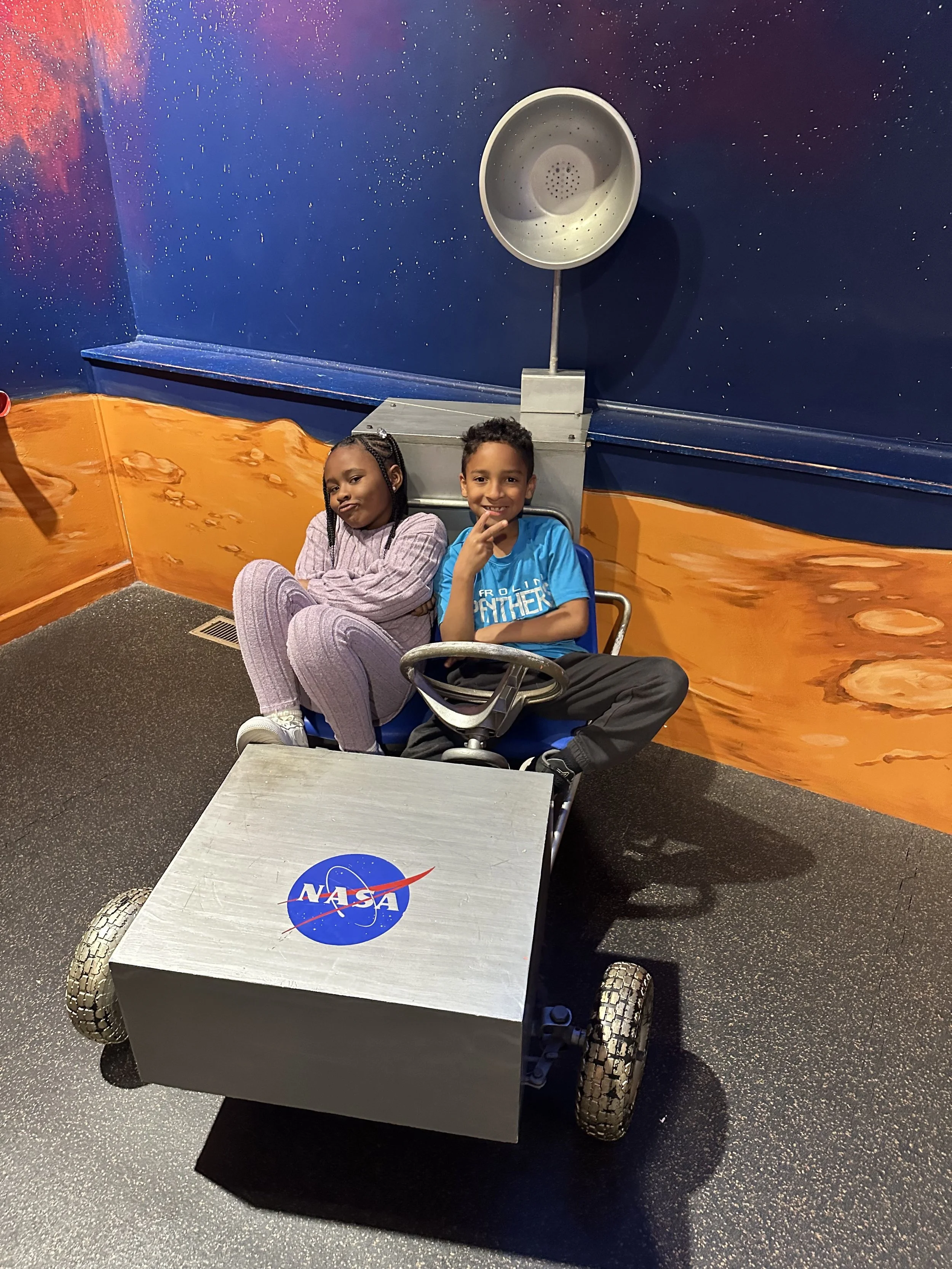 Two children sitting in a NASA-themed space rover mock-up, with a background painted to resemble space and planets.