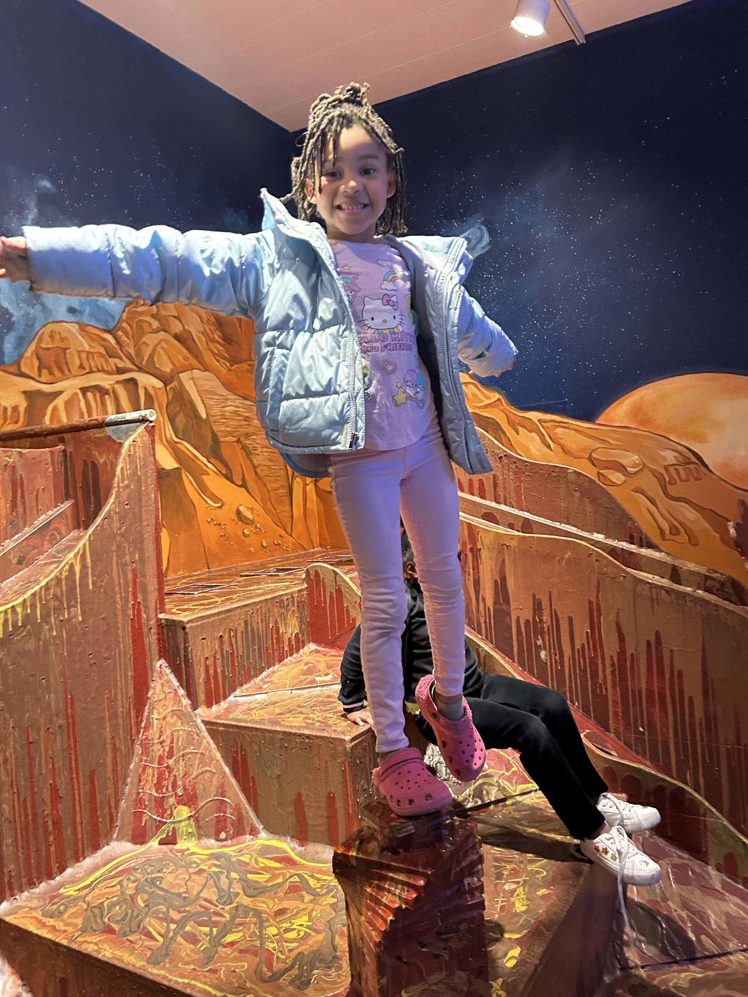 A young girl with dreadlocks smiling and extending her arms, standing on a colorful, art-designed staircase with a mural of mountains and a starry night sky in the background.