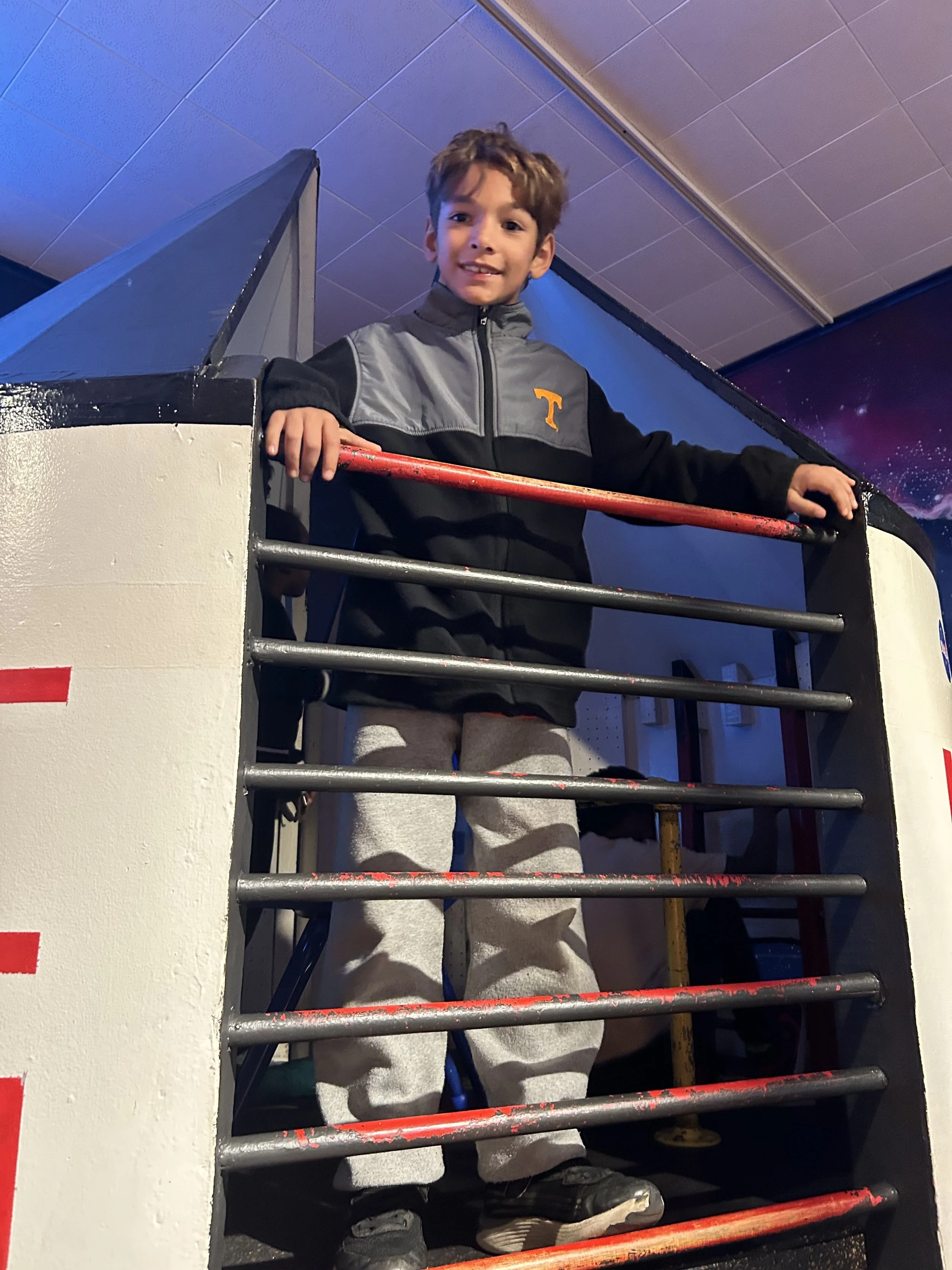 Young boy standing in a space-themed exhibit, holding onto metal bars, wearing a black and gray jacket with a Tennessee Volunteers logo, gray sweatpants, and black sneakers.