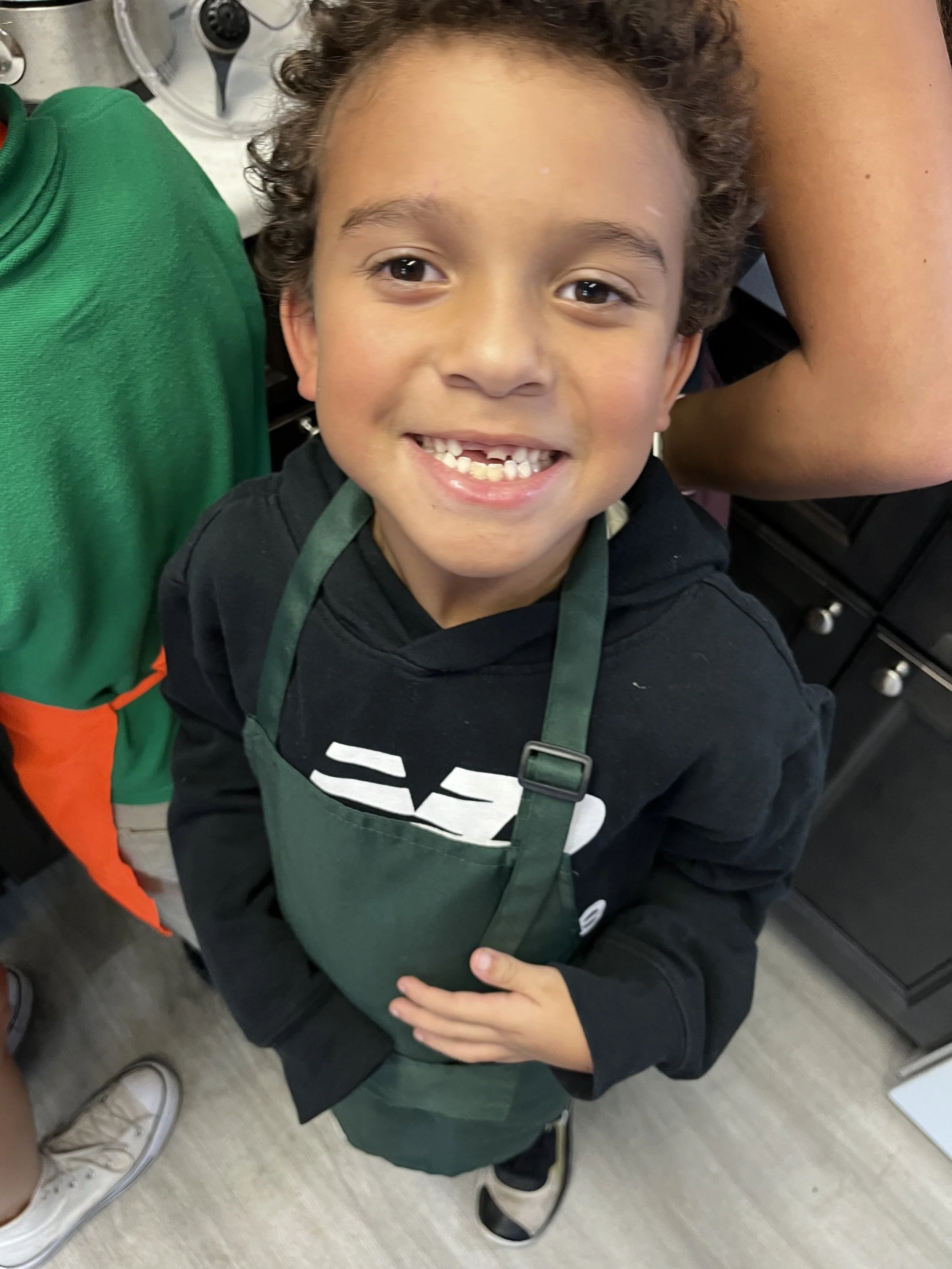 A young boy with curly hair, wearing a black hoodie and a green apron, smiles at the camera with missing front teeth. He is standing indoors, surrounded by people in an environment that looks like a kitchen or classroom.