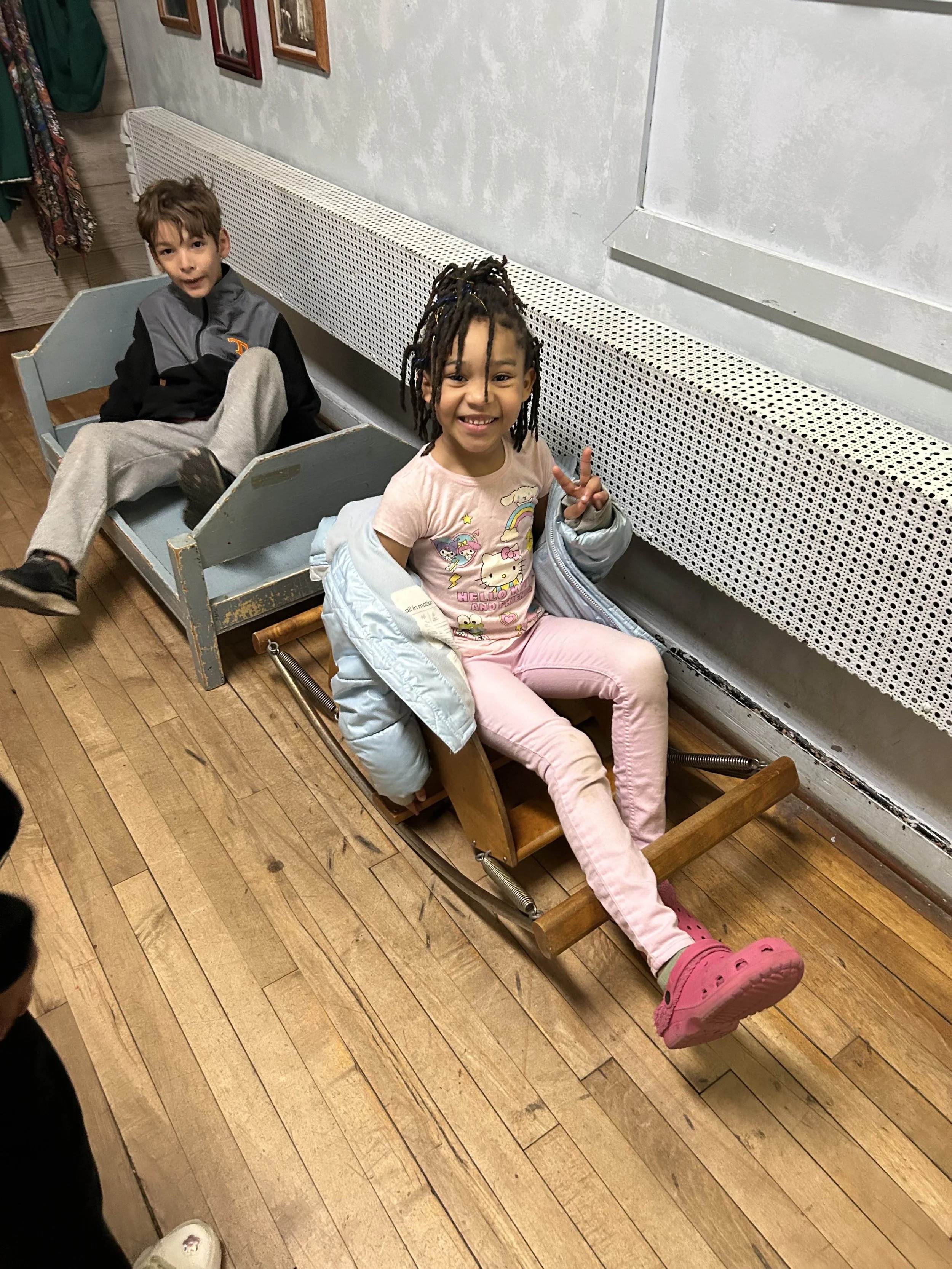 Two children sitting on vintage wooden sleds indoors, with wooden flooring and a wall with framed pictures in the background.