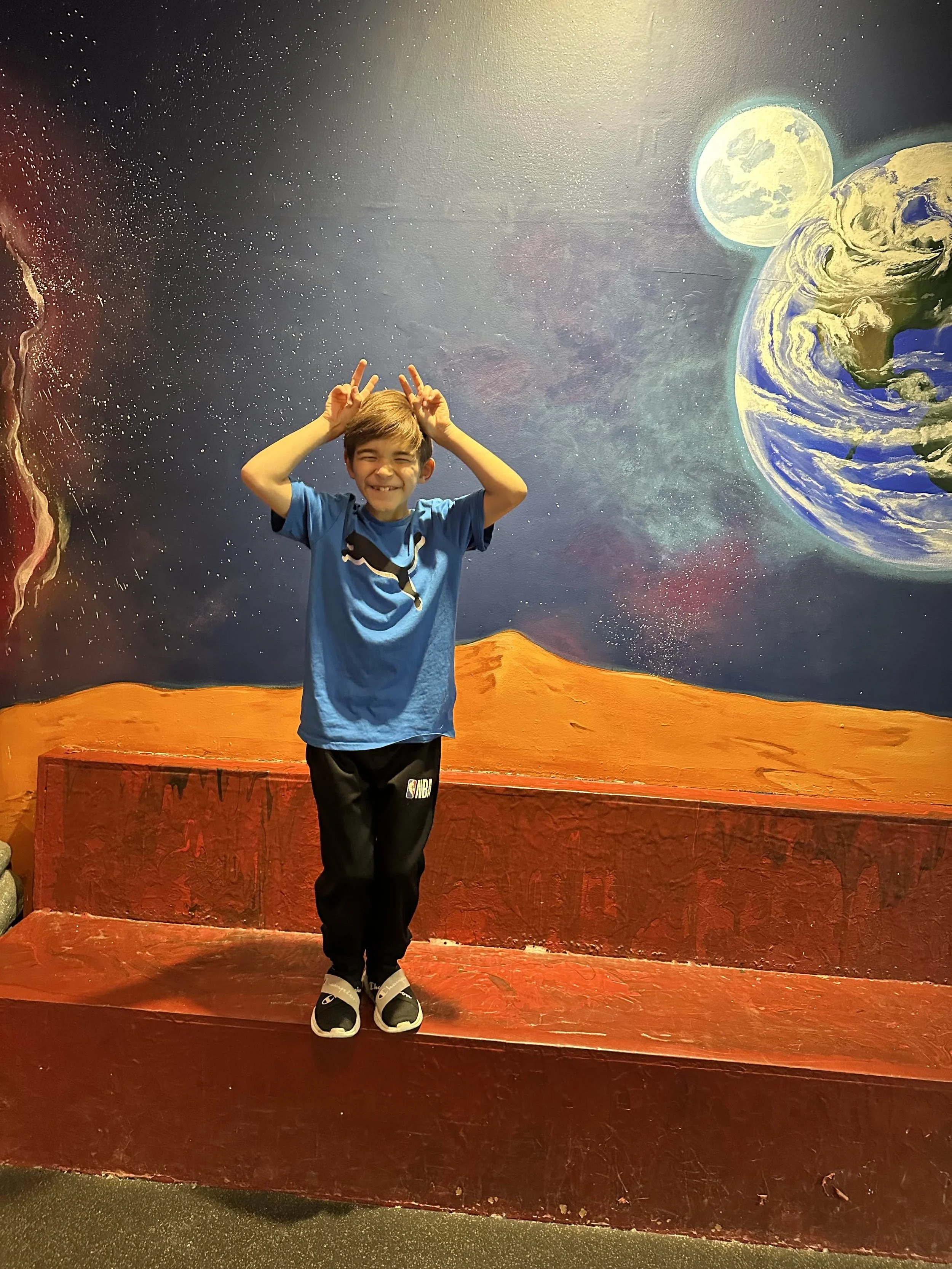 A smiling boy in a blue shirt with a graphic design, posing with peace signs behind his head, standing on a painted red platform with a space-themed mural in the background showing planets, stars, and celestial bodies.
