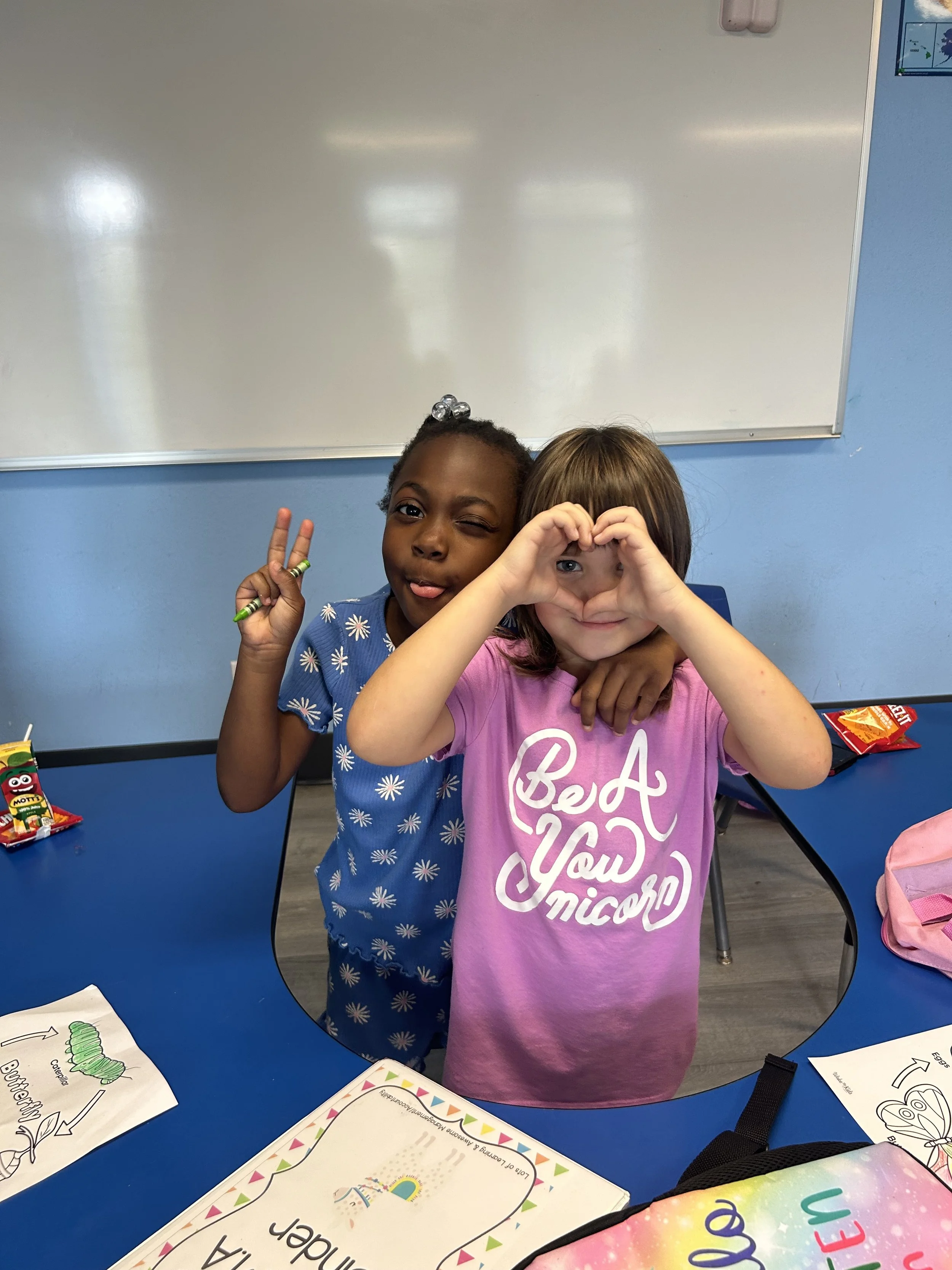 Two young girls stand at a blue table in a classroom, one making a peace sign, the other forming a heart shape with her hands around her eyes. There are various school supplies and worksheets on the table.