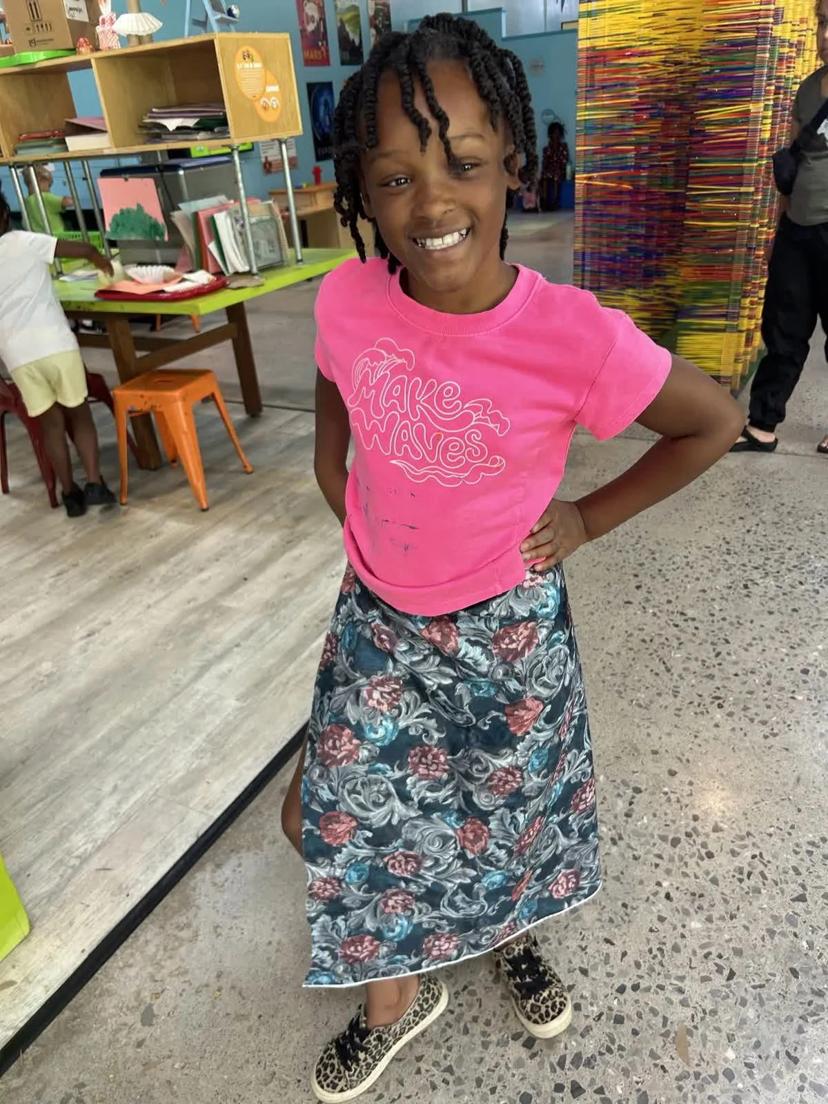 A smiling young girl with curly hair, wearing a pink t-shirt and a floral skirt, stands with her hand on her hip in a colorful indoor setting that appears to be a play area or classroom.