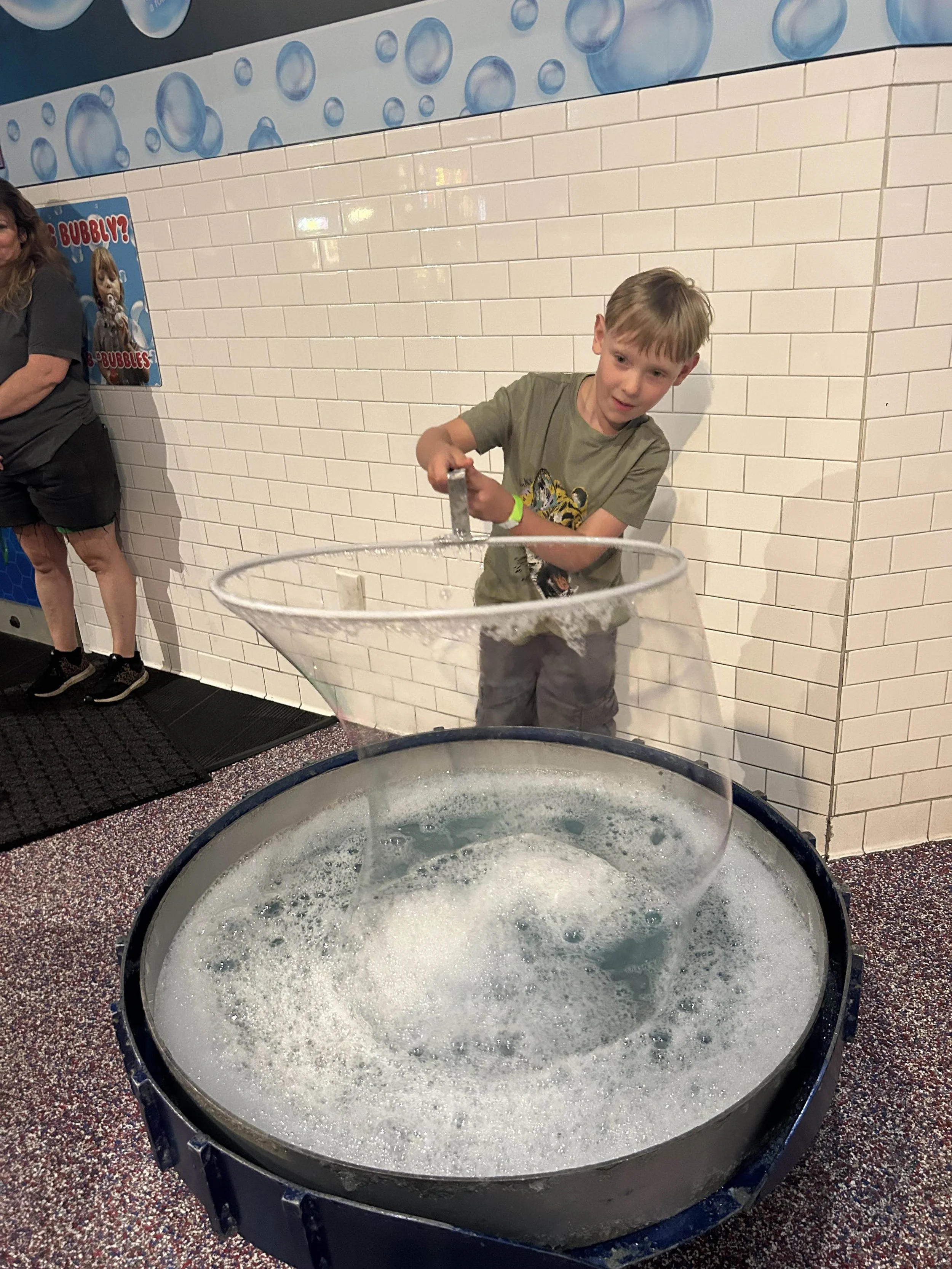 A young boy is using a hammer and chisel to break a large bubble into a smaller one, at an indoor science or amusement center with tiled walls and foam or bubbles in a large container.