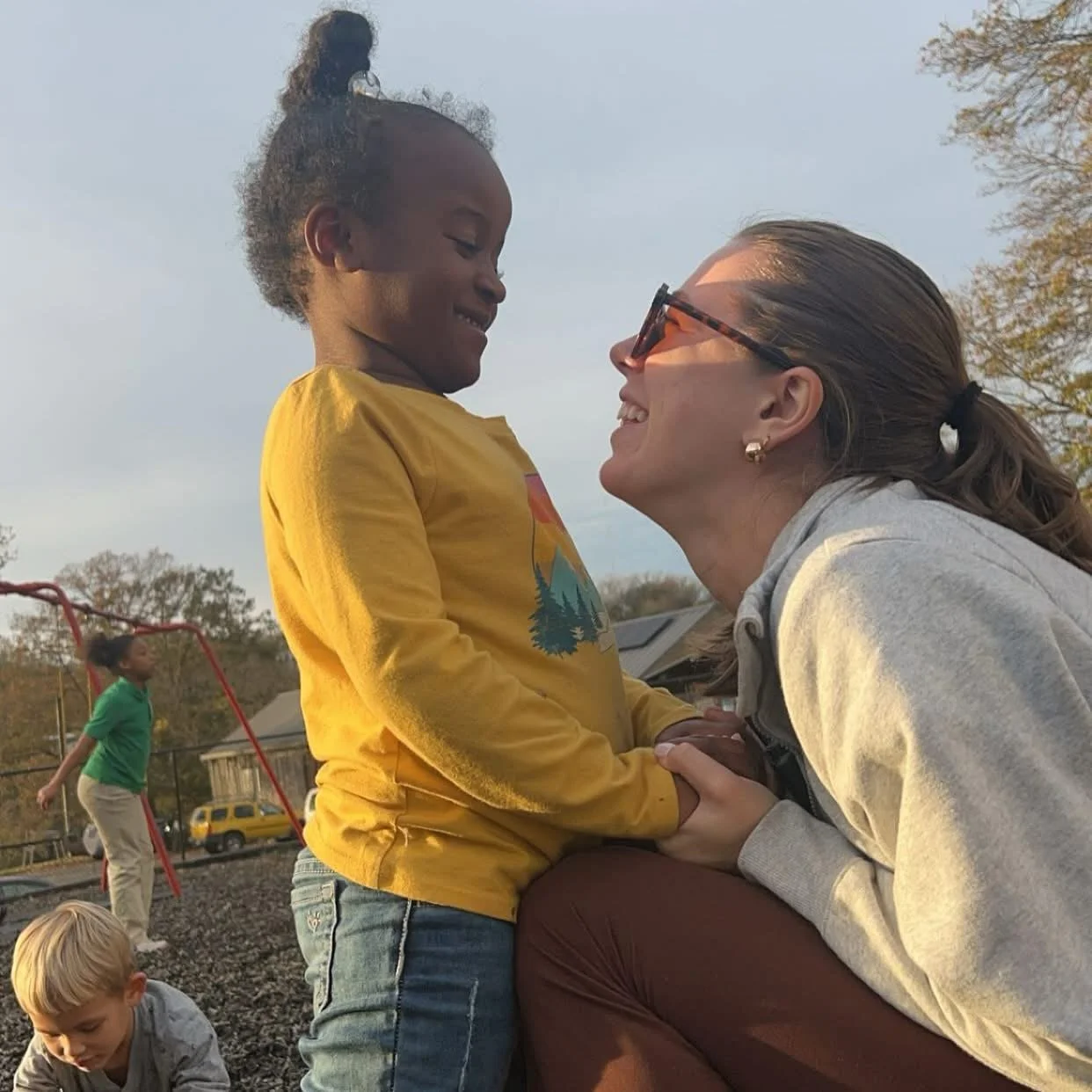A woman and a young girl are smiling at each other at a playground, with other children playing in the background.