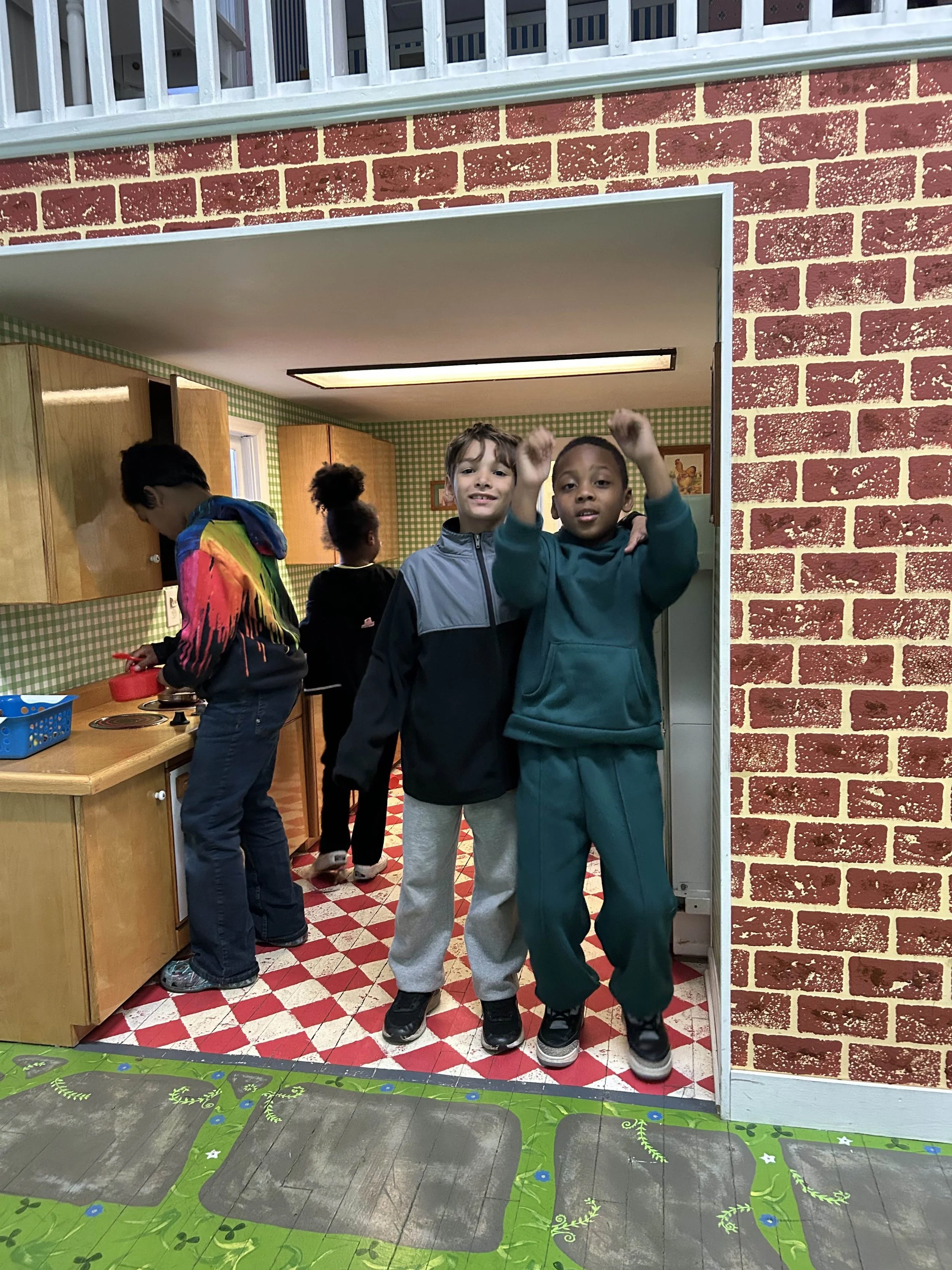 Two boys standing in front of a doorway, smiling and raising their arms inside a colorful kitchen with children in the background.