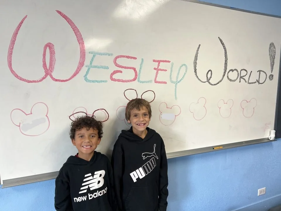 Two young boys standing in front of a whiteboard with colorful writing and drawings. The text says 'QWEEN SEO WORLD!' with various doodles of bear head shapes around.