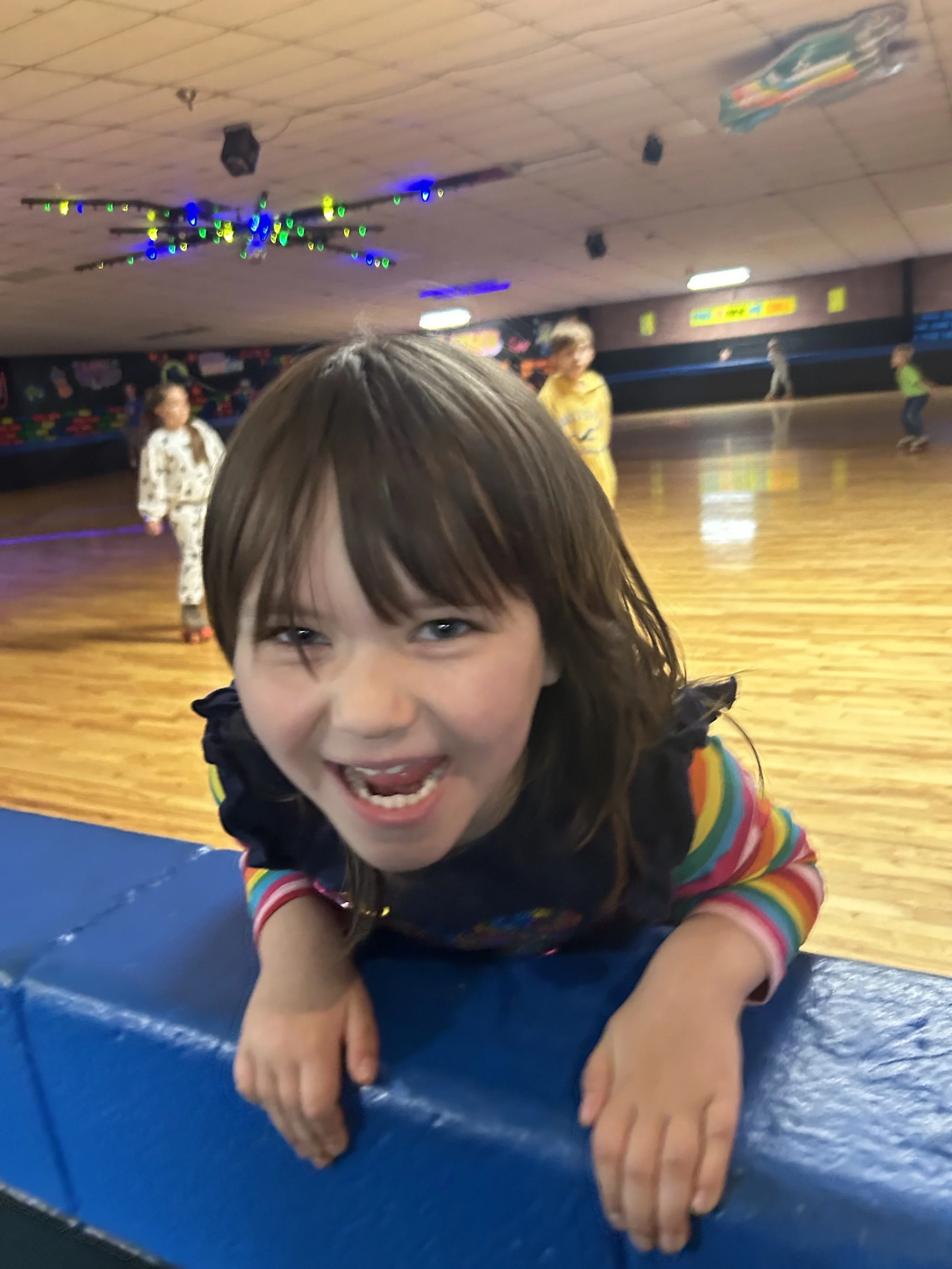 Young girl with brown hair and striped rainbow shirt smiling and leaning over a blue padded surface in a roller skating rink, with other kids skating in the background.
