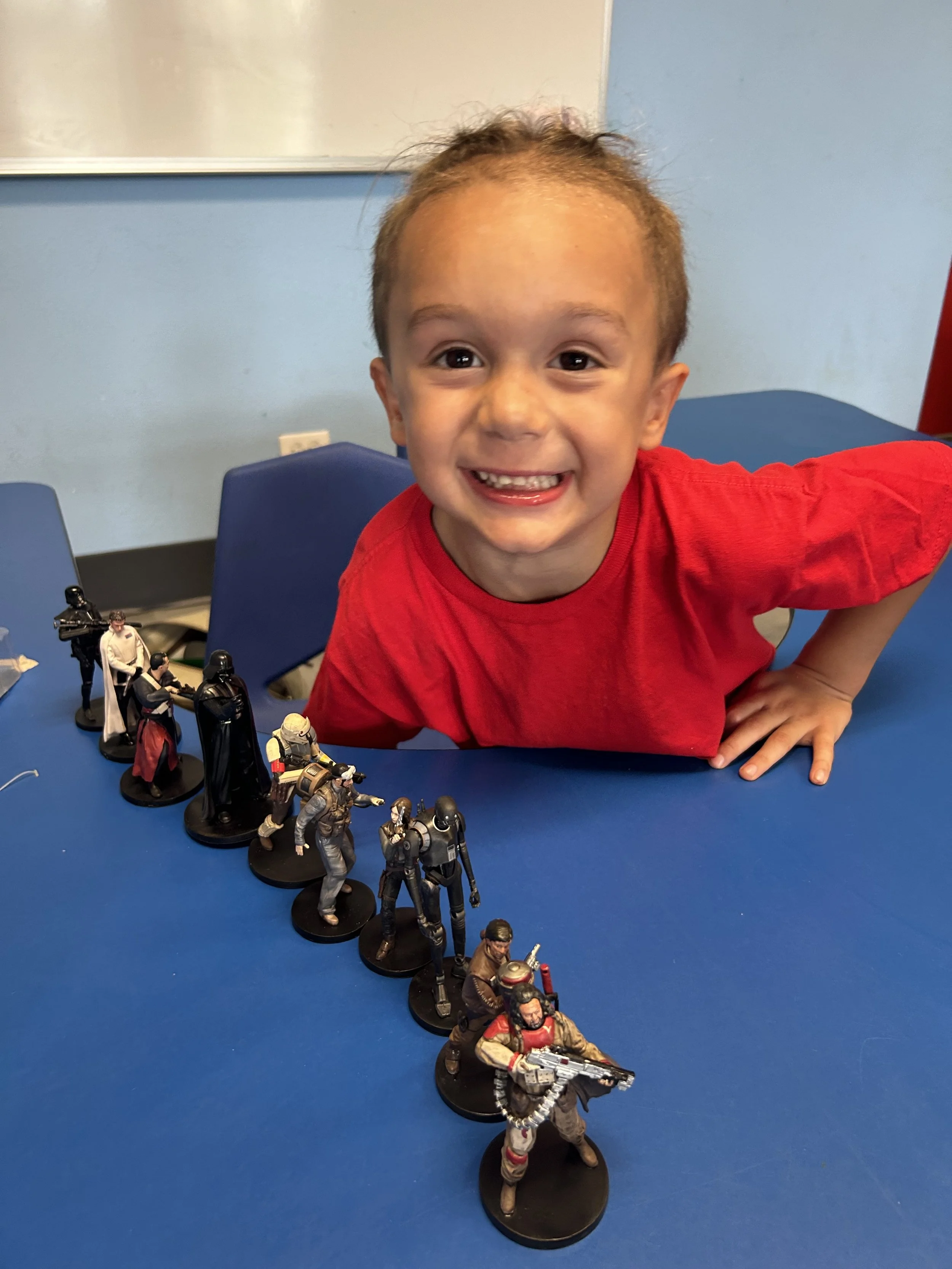 A young boy with curly hair, wearing a red shirt, smiling and leaning on a blue table. In front of him are various Star Wars figurines arranged in a line.