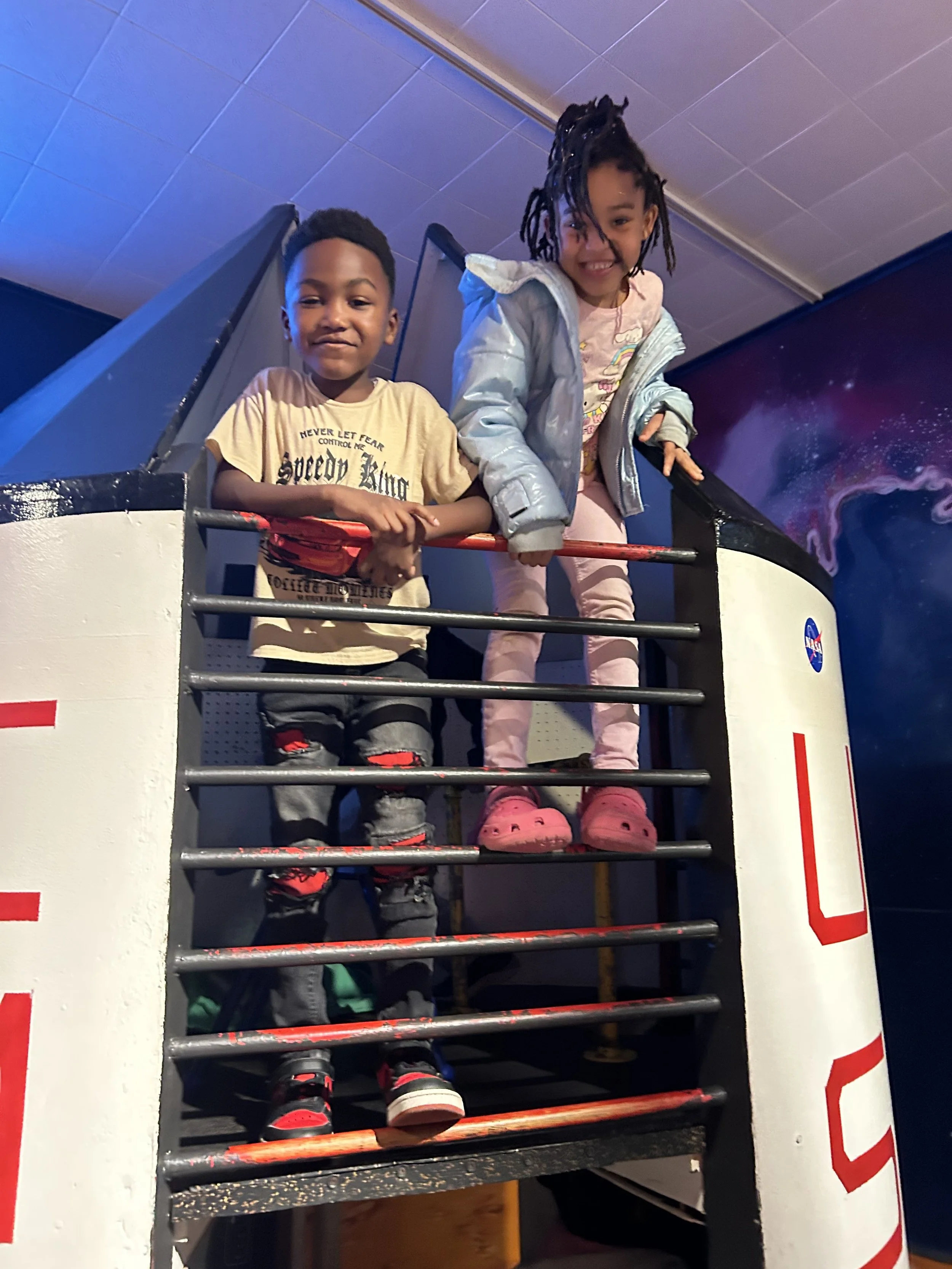 Two children standing on the metal stairs of a spaceship exhibit at a science museum, smiling at the camera.