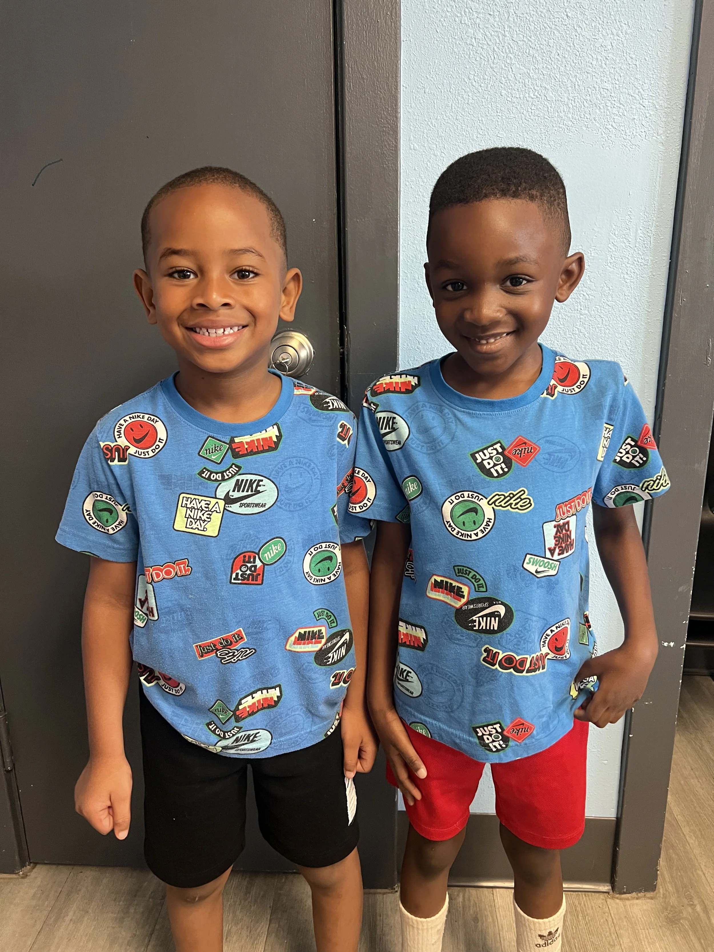 Two young boys standing side by side, smiling, wearing matching blue Nike t-shirts with various logos and slogans, one in black shorts and the other in red shorts, inside a building near a dark locker.