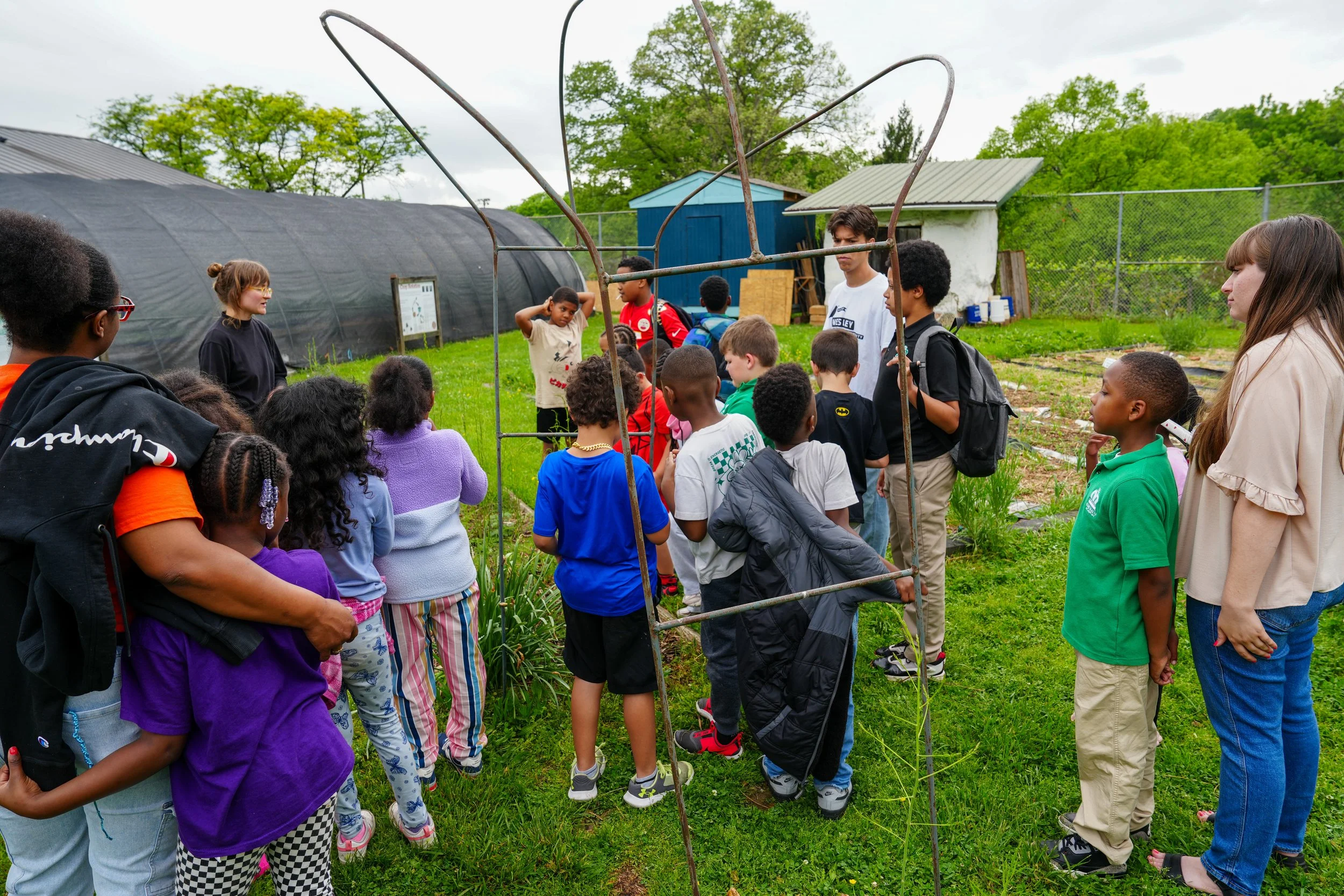 A group of children and adults gathered outdoors in a garden or farm setting, listening to a woman speak near a small structure made of metal rods, with greenery and sheds in the background.