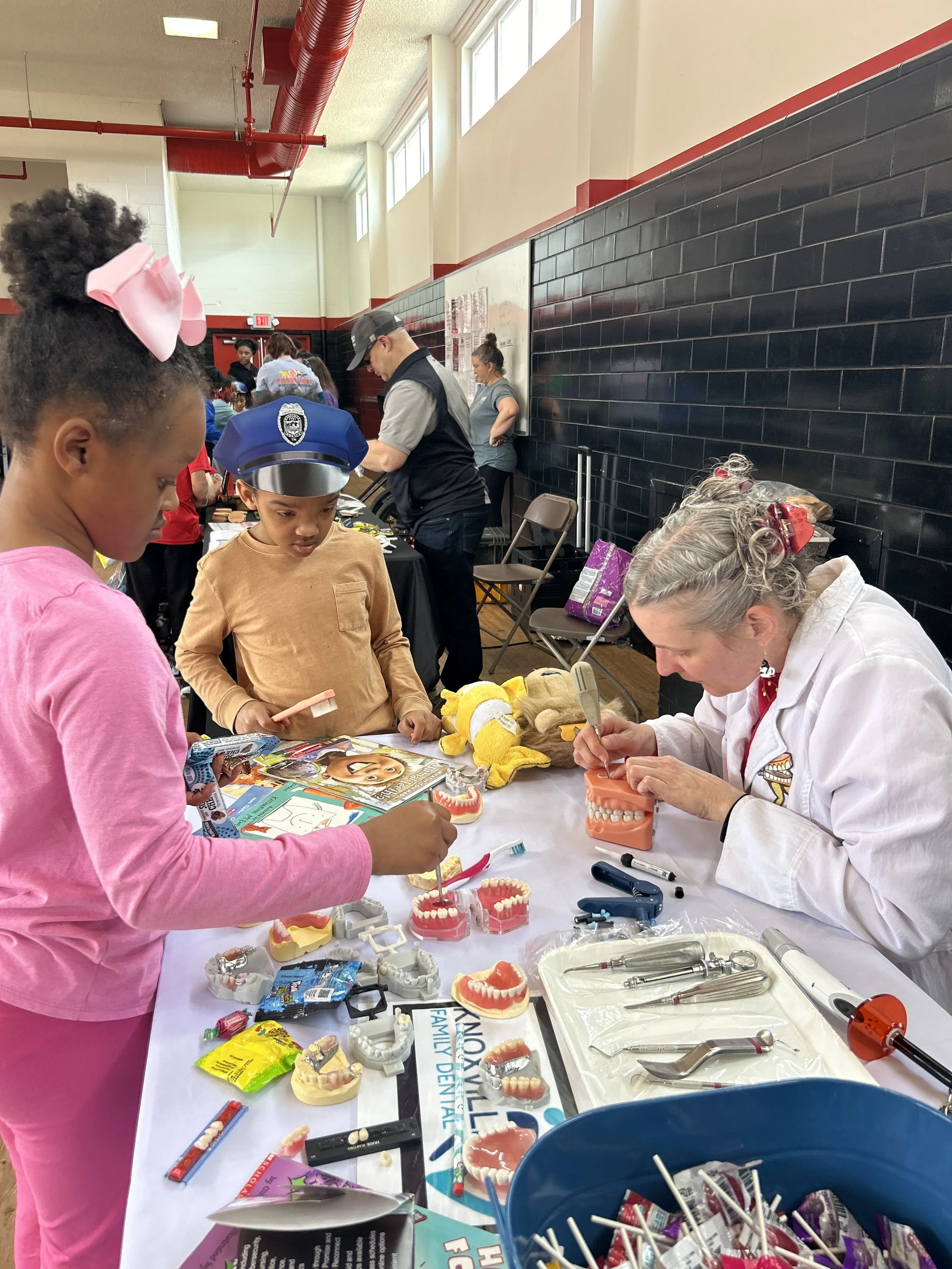 Children exploring dental health education with a dentist, who is showing models of teeth and dental tools on a table at a community event.
