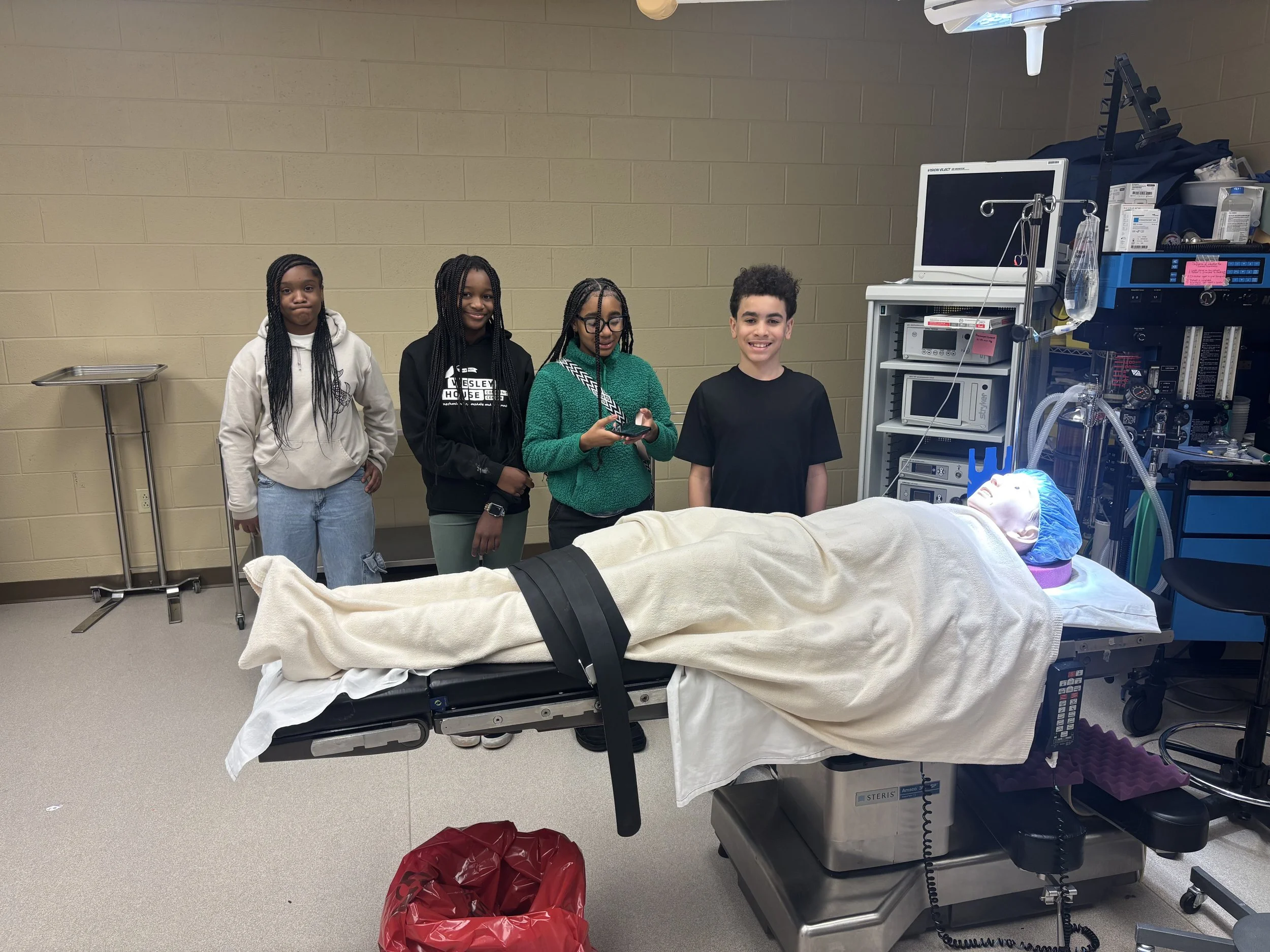 Four children stand behind a hospital gurney with a medical mannequin head and torso, in a clinical room with medical equipment.