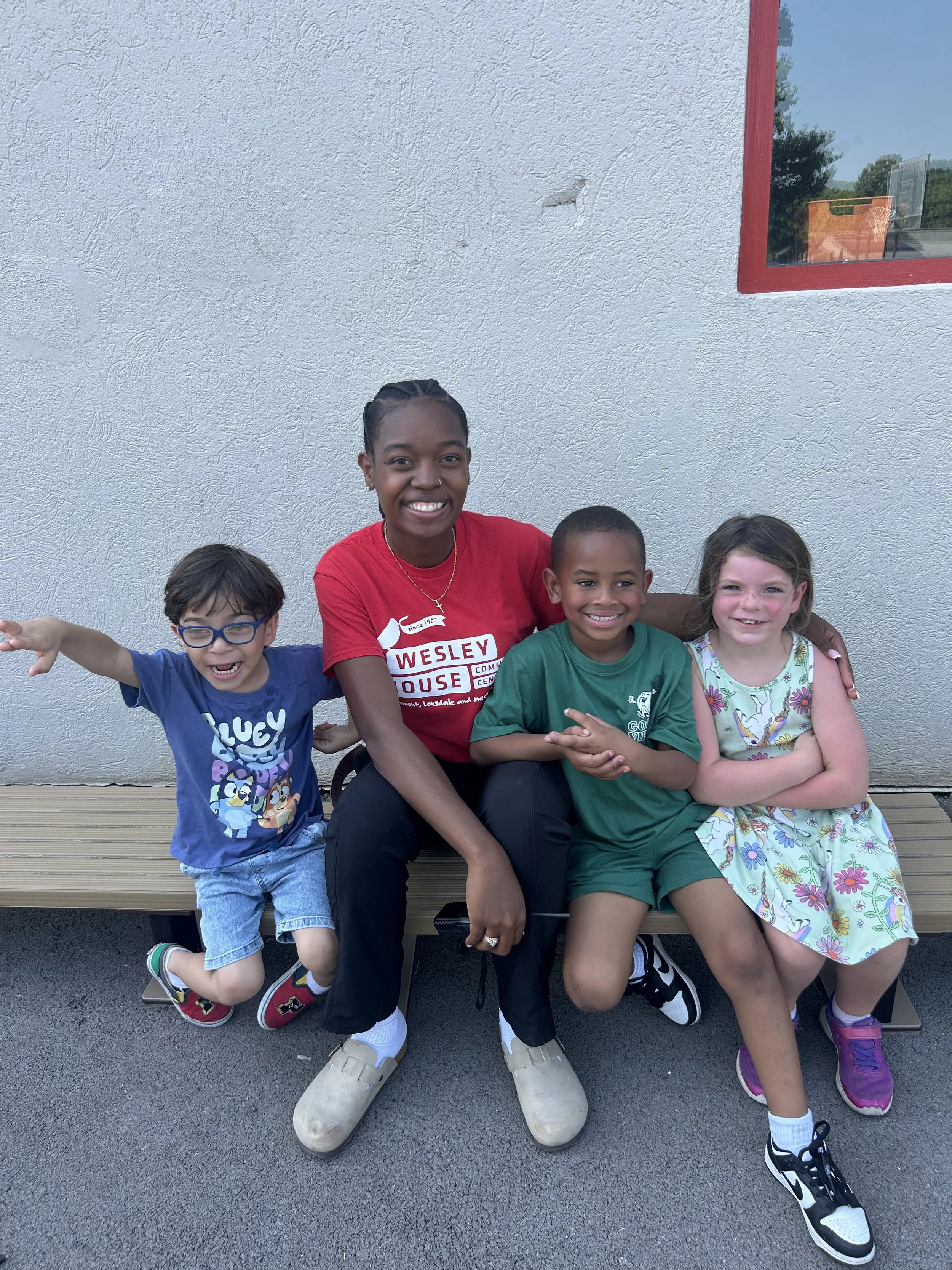A woman and three children sitting on a bench outside against a plain wall. The woman is smiling and wearing a red shirt, with three children sitting beside her, all smiling and looking at the camera.