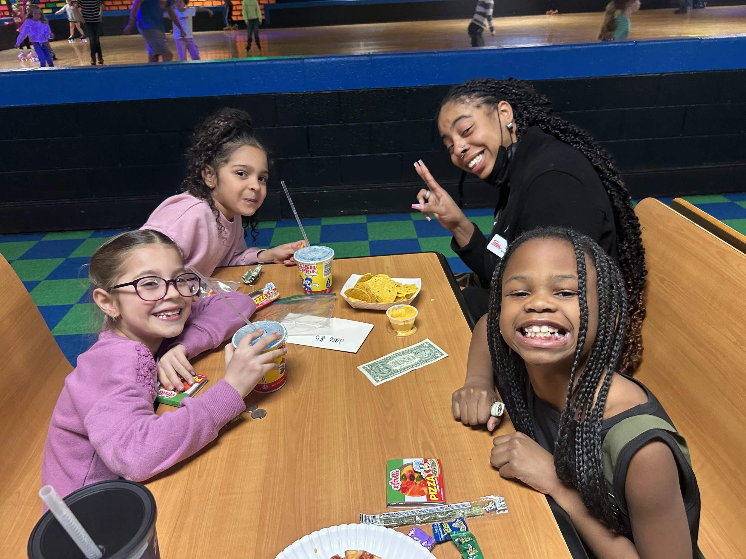 Four young girls and an adult woman sitting at a table in a roller skating rink, enjoying snacks and drinks, smiling at the camera.