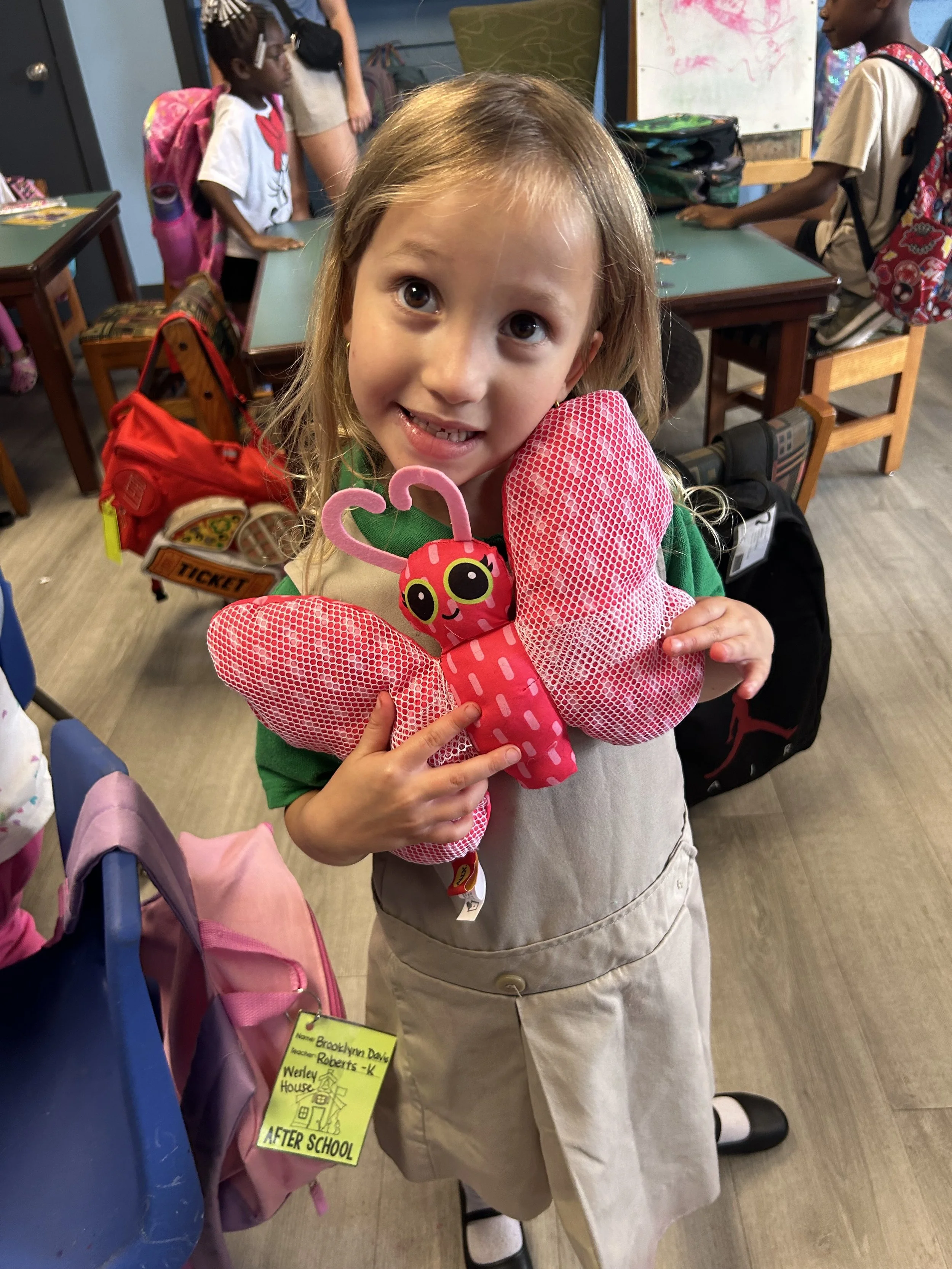 A young girl with blonde hair holding a pink butterfly-shaped backpack with a ladybug face, smiling at the camera in a classroom with other children and backpacks in the background.