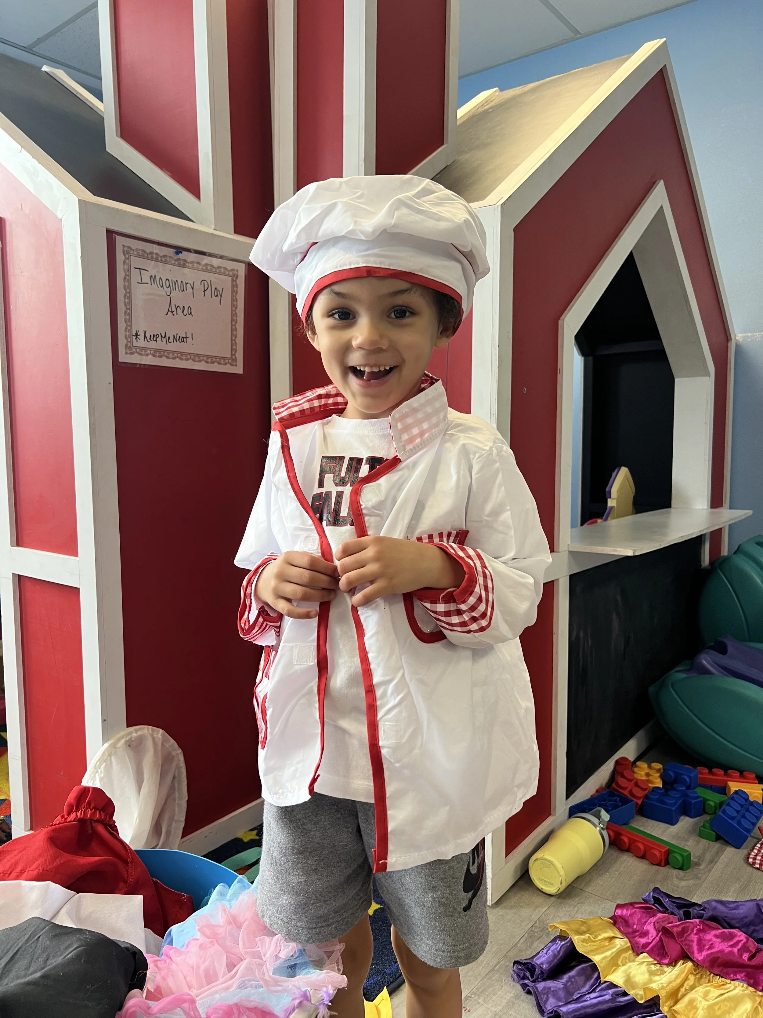 Child dressed as a chef standing in front of a playhouse in an imaginary play area, surrounded by toys and colorful outfits.
